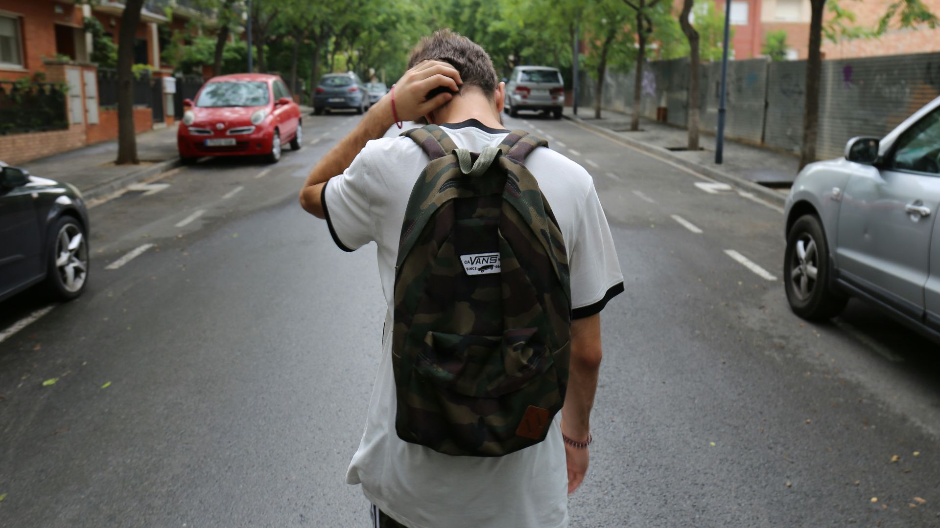boy wearing white shirt and black shorts carrying backpack standing on black concrete road between vehicles and trees during daytime