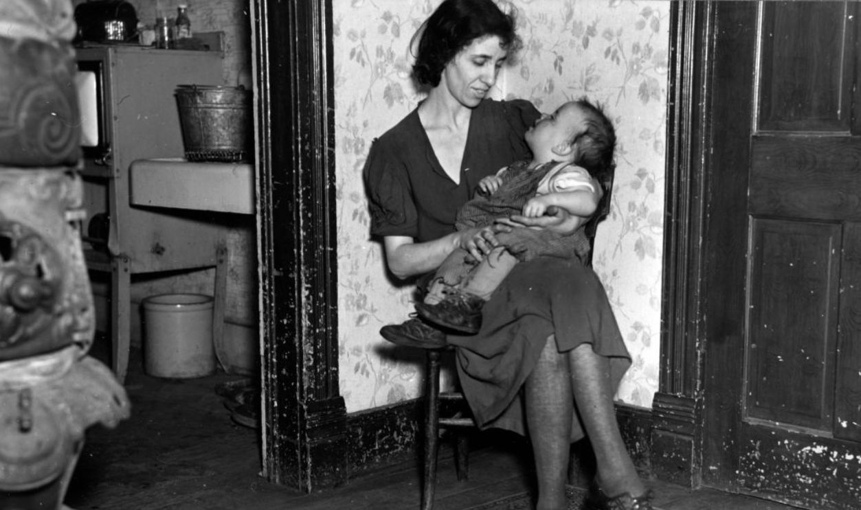 A woman, Mrs Homer Sharer, holds her baby in her lap while seated in a room, Estherville, Iowa, December 1936. The Sharers are former tenant farmers but are living on unemployment relief. 