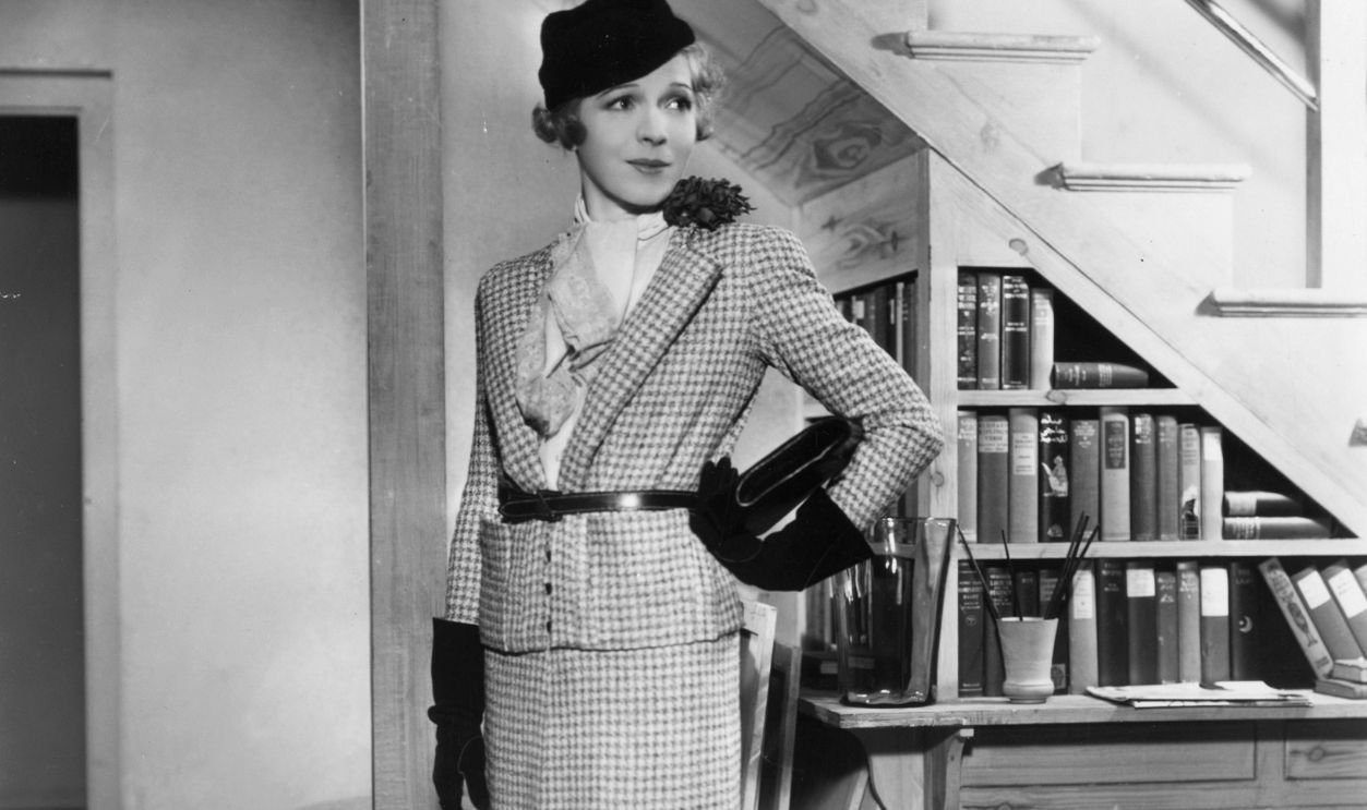 27th April 1934: An actress waits somewhat haughtily in a hallway wearing a yellow twill gabardine suit and carries a clutchbag on the set of 'Biography' staged at London's Globe Theatre.