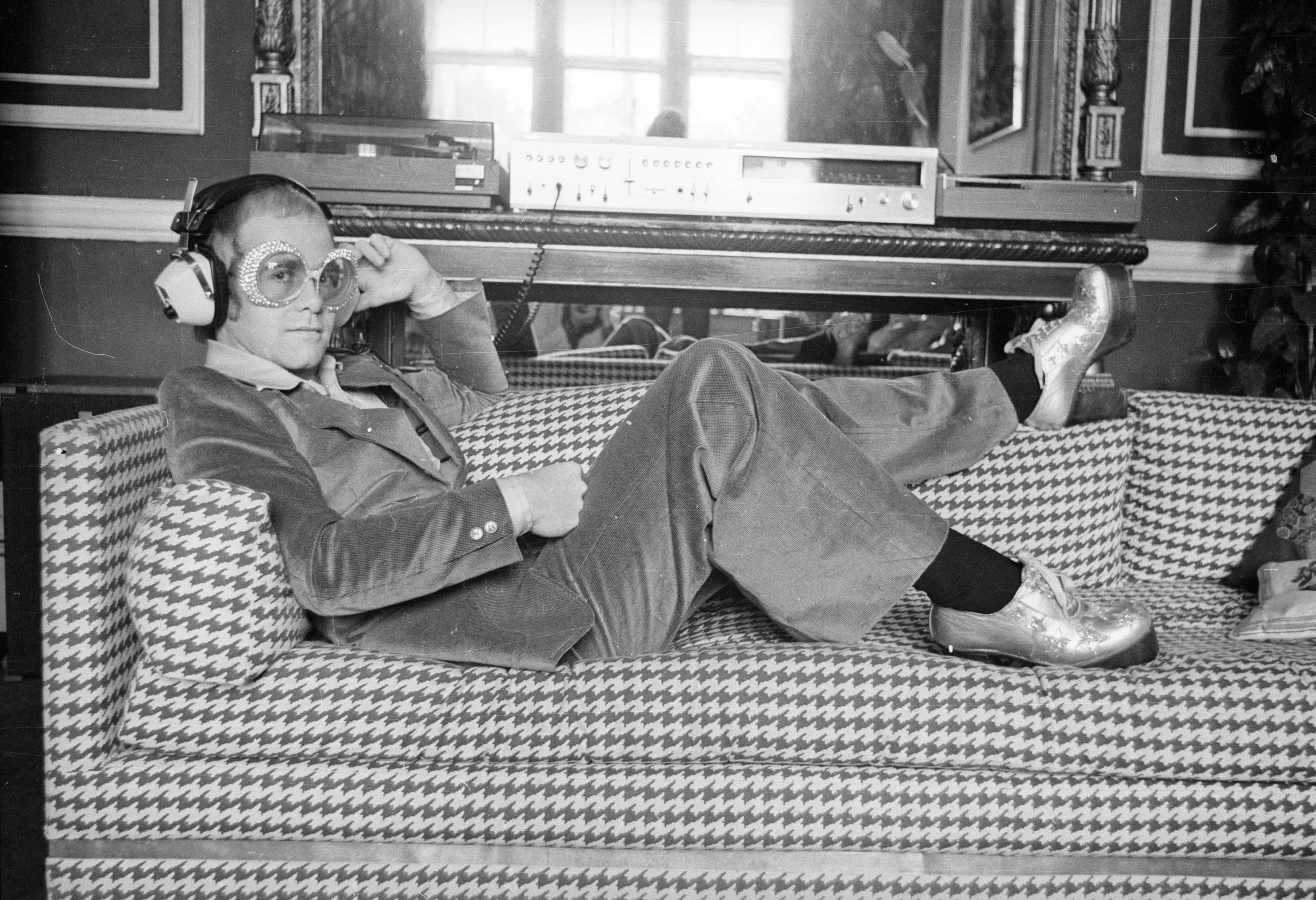 Elton John relaxes on a sofa and listens to some music - 1974