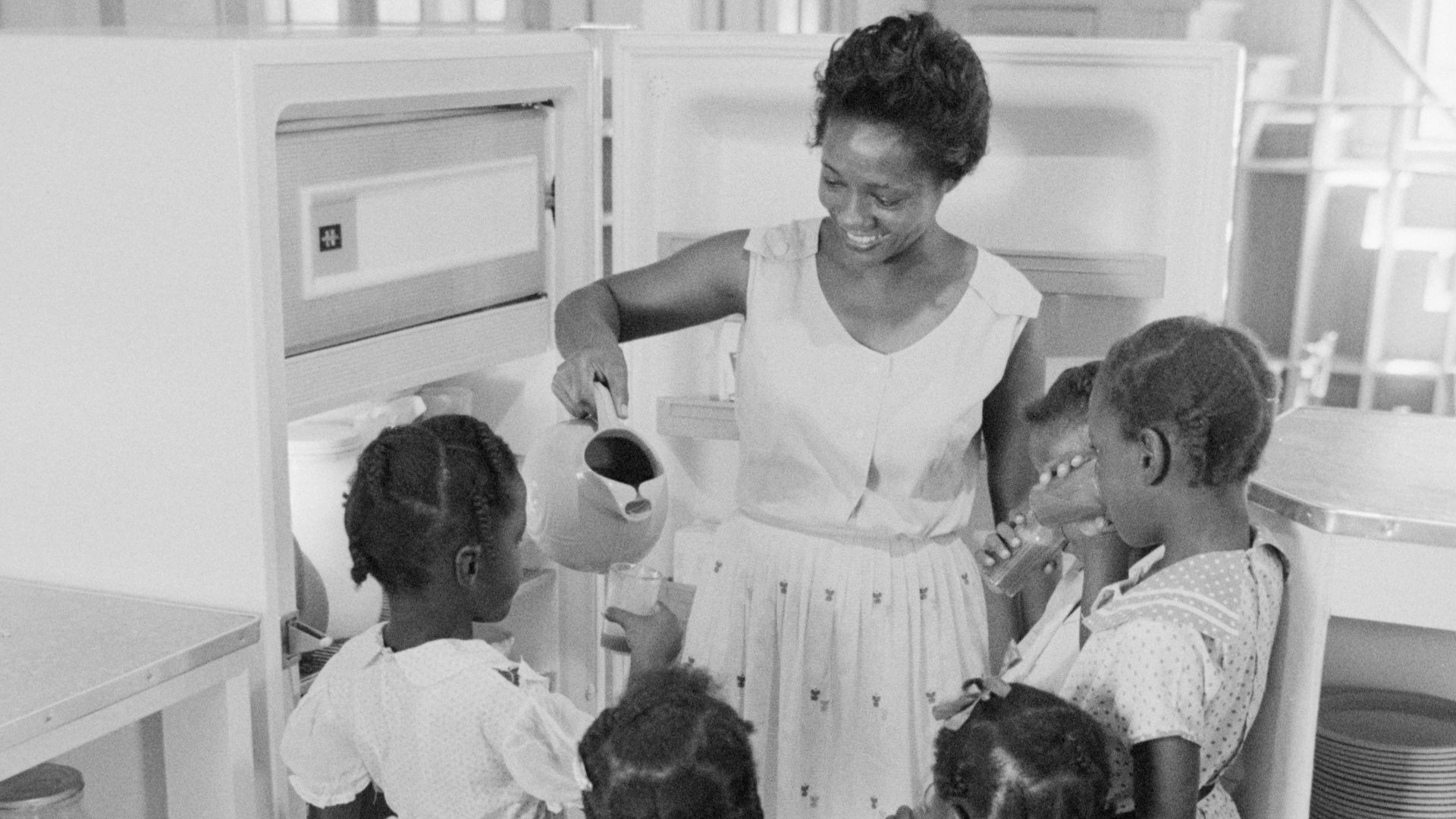 African American woman at refrigerator holding a pitcher as she pours out drinks for children, probably Little Rock, Arkansas