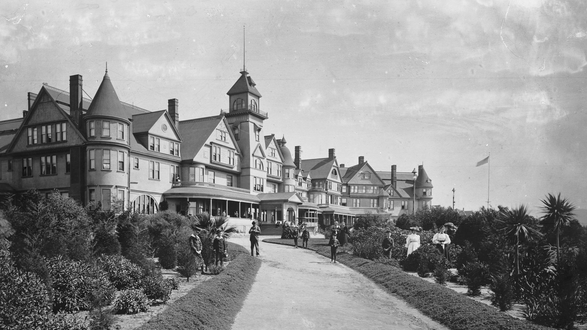 The Hotel Redondo, ca.1900
Photograph of the Redondo Hotel and gardens, ca.1900. Looking southeast up a long walkway to the hotel. Several people are standing in the gardens and on the path. The majestic three-story hotel is Victorian in design. A flag is