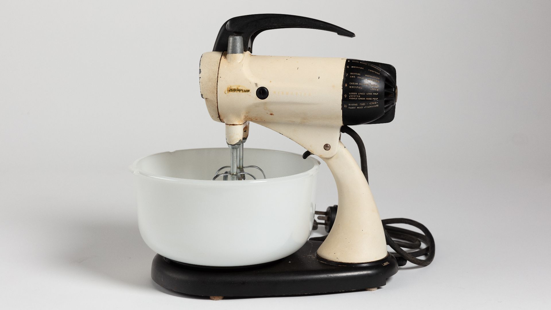 Sunbeam Mixmaster cake mixer. Cream metal with some black bakelite/plastic parts. Two opaque glass bowls. Two metal mixing arms. Collection of Auckland Museum Tāmaki Paenga Hira, 2023.43.2, gift of Janene Smith.