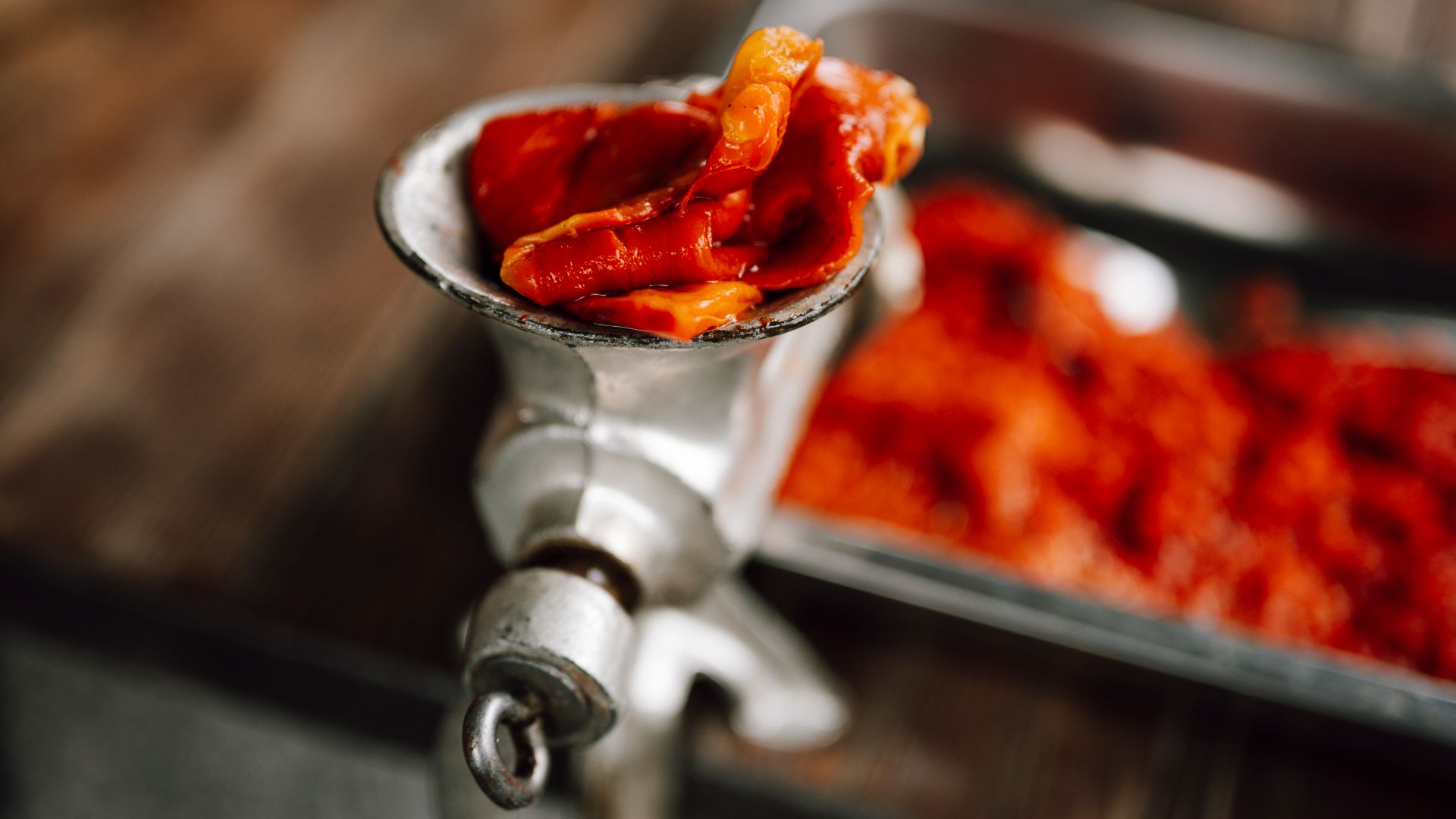 Fresh, ripe tomatoes are being processed in a metal food grinder. The wooden table shows other prepared ingredients, adding a homey feel to the cooking activity.