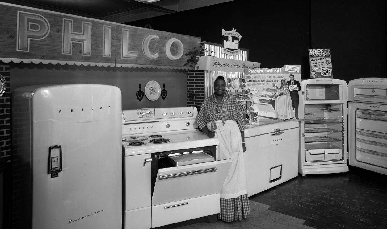 Woman dressed as Aunt Jemima standing with merchandise display, at the Casey and O'Brien Store, 124 West Mifflin Street, featuring Philco appliances including two refrigerators, a chest freezer, and a stove, Madison, Wisconsin, February 27, 1954.