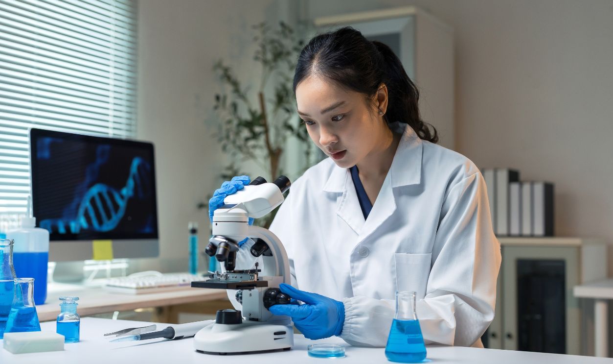 Asian female scientist wearing lab coat and blue gloves, studying biological sample with microscope in a modern research lab
