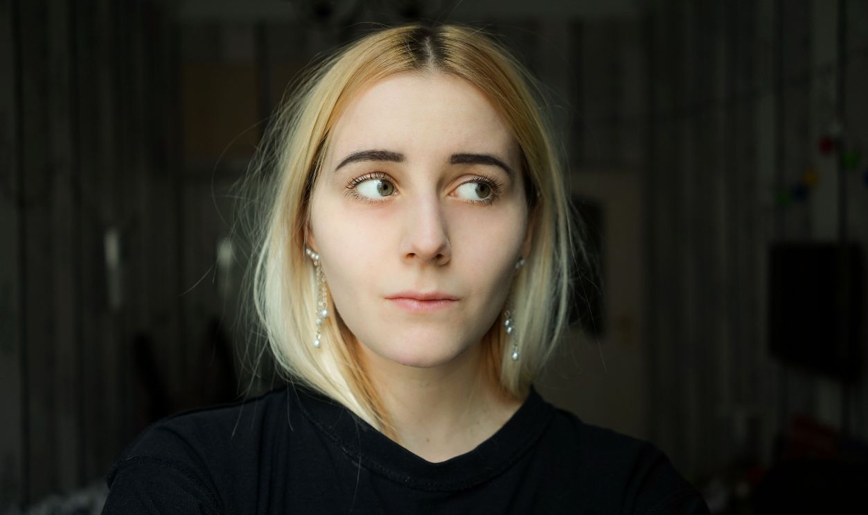 A thoughtful young woman with blond hair looks to the side indoors, reflecting quietly.