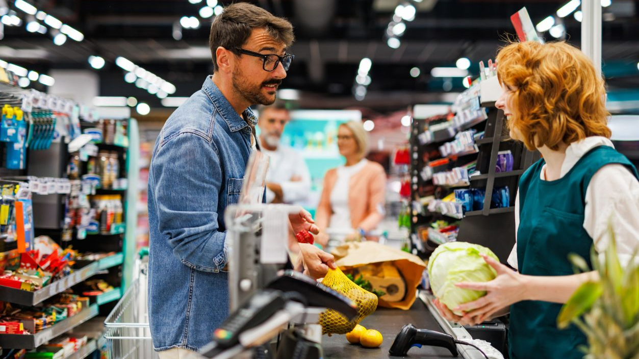 Shutterstock-2546757641, Supermarket cashier scanning groceries at the checkout counter while talking with a male customer, other customers waiting in line behind