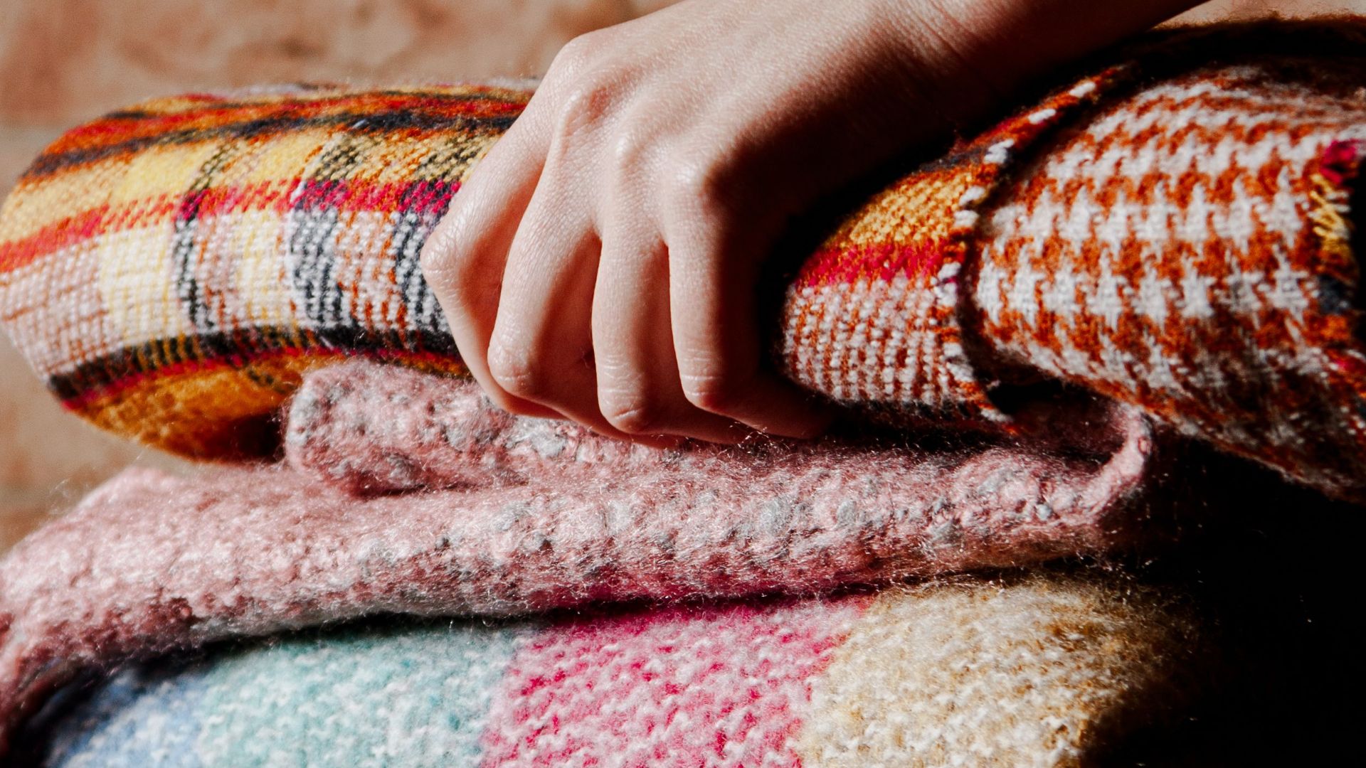 Hand gently touching a stack of colorful textured wool blankets in warm light.