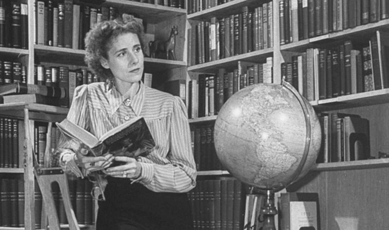 Author Clare Boothe Luce at home in her library, wearing pants, leaning on library ladder near globe and holding open a copy of a biography of Madame Curie; this photograph is autographed by Mrs. Luce.