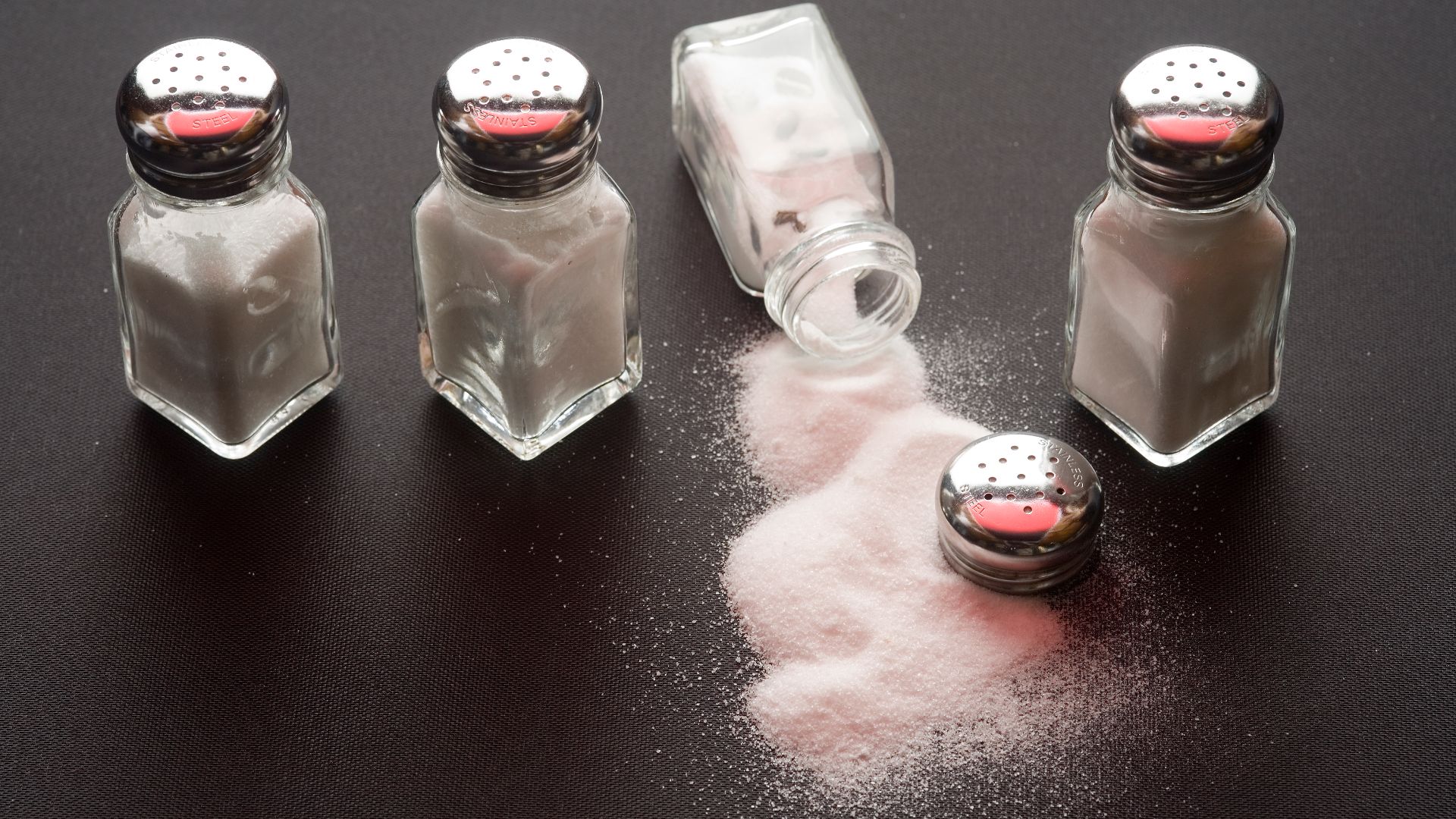 Four salt shakers on a table with salt spilled from one of them.