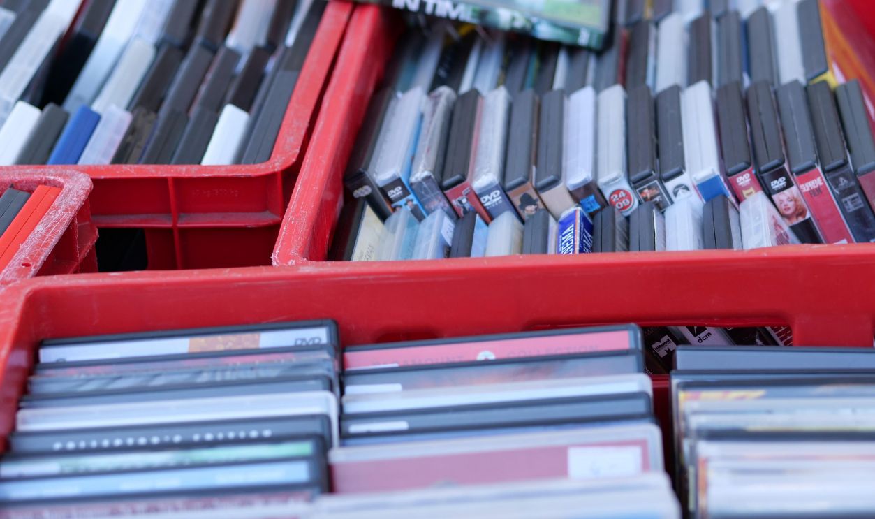  Dusseldorf, North Rhine-Westphalia Germany – March 22 2026 A high-angle close-up of several bright red plastic crates overflowing with a vast collection of second-hand DVDs and Blu-rays at an outdoor flea market. The movie cases, including titles like "In Time," are tightly packed, showing a mix of black, white, and blue spines. The bright morning sunlight highlights the repetitive patterns of the boxes, capturing the essence of physical media nostalgia and urban bargain hunting.
