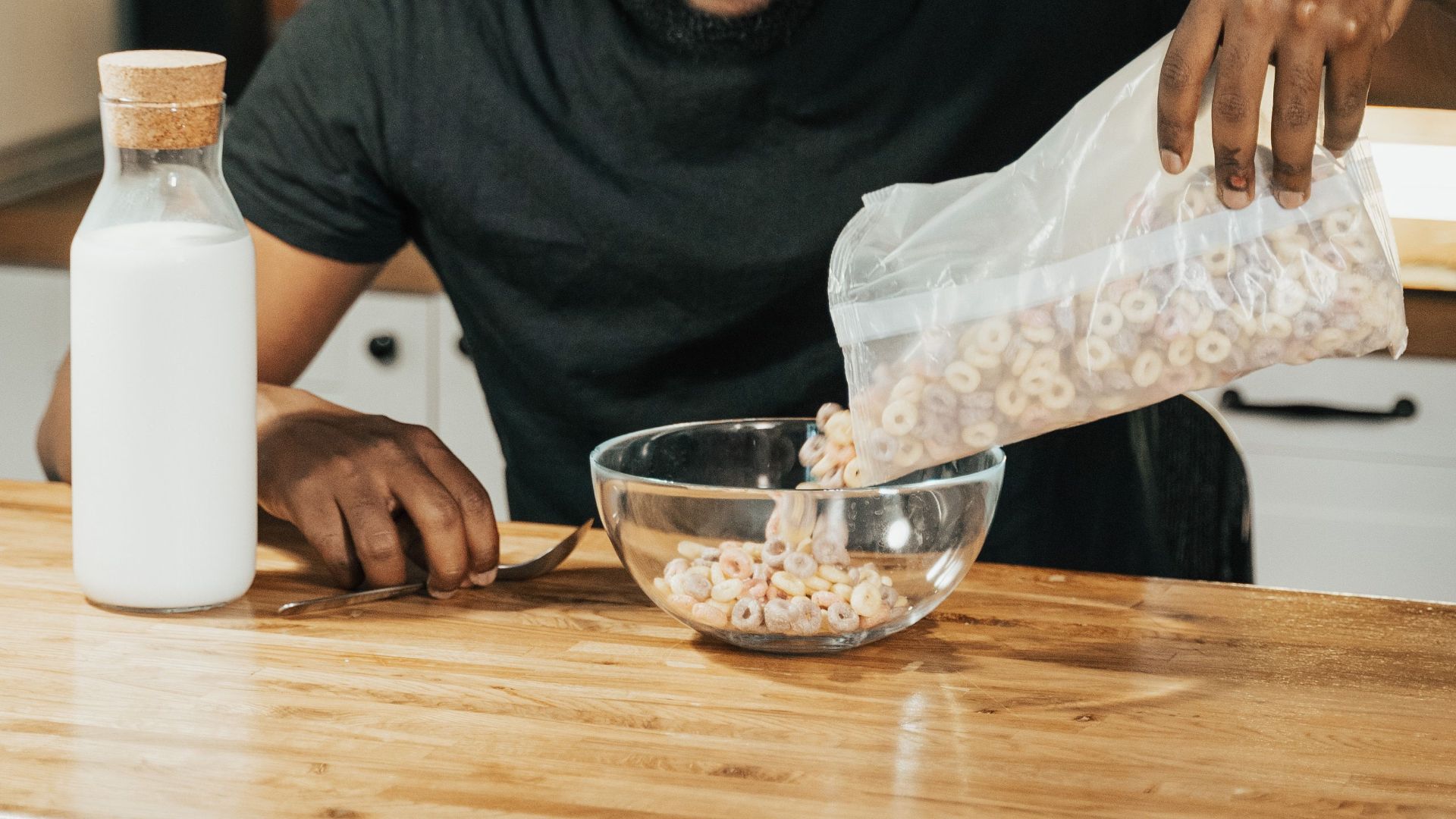 Man pours cereal into a bowl for breakfast at a wooden table indoors.