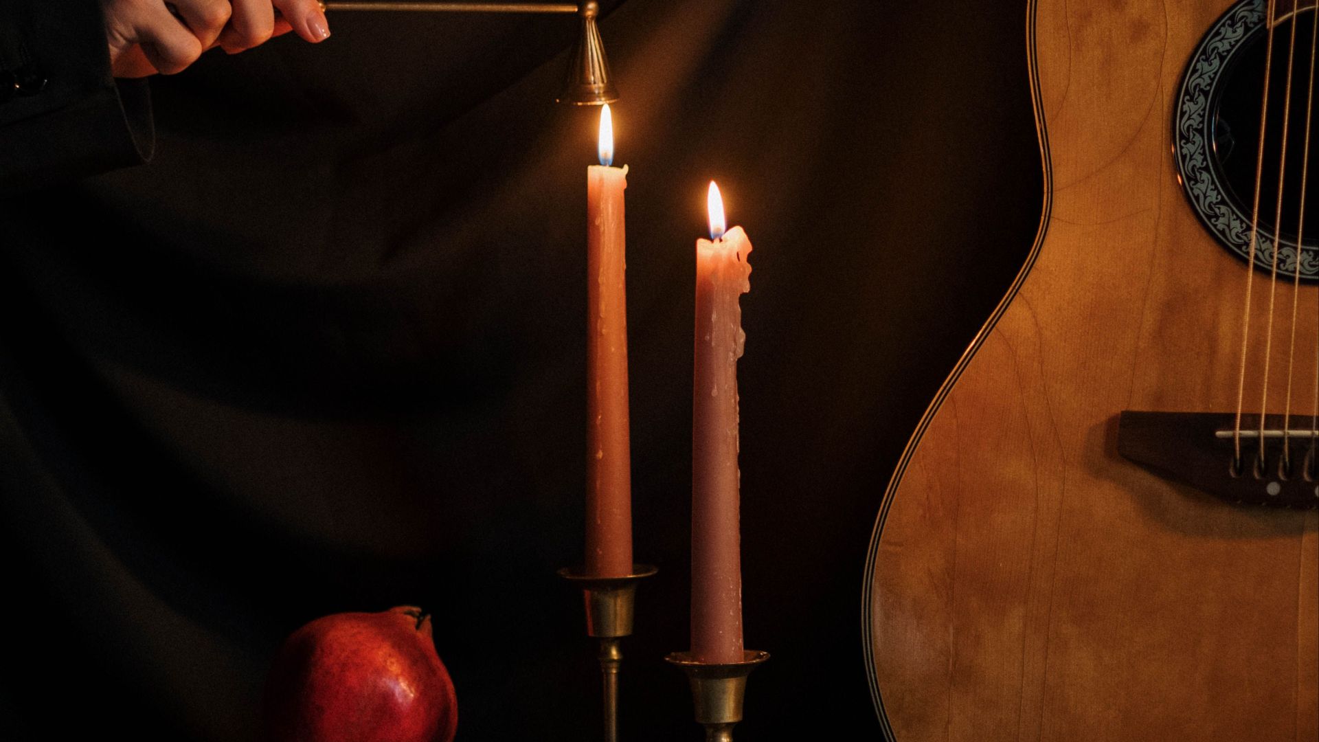 A dark still life scene with candles, a guitar, books, and a pomegranate, evoking a vintage mood.