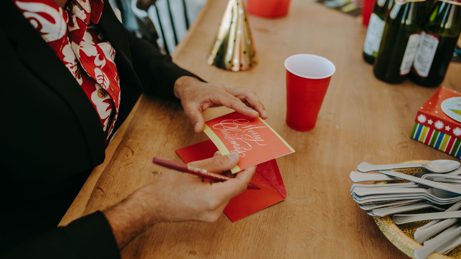 Person writing a Merry Christmas card at a festive table setting with drinks and utensils.