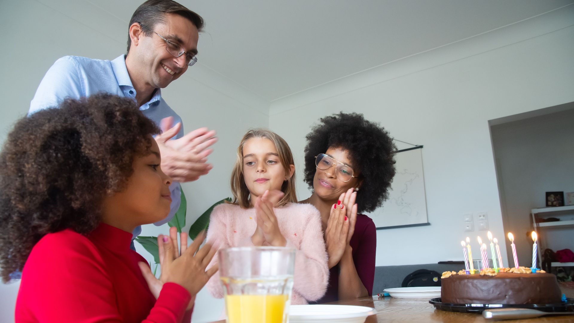 A diverse family clapping and celebrating a birthday with a chocolate cake and candles indoors.