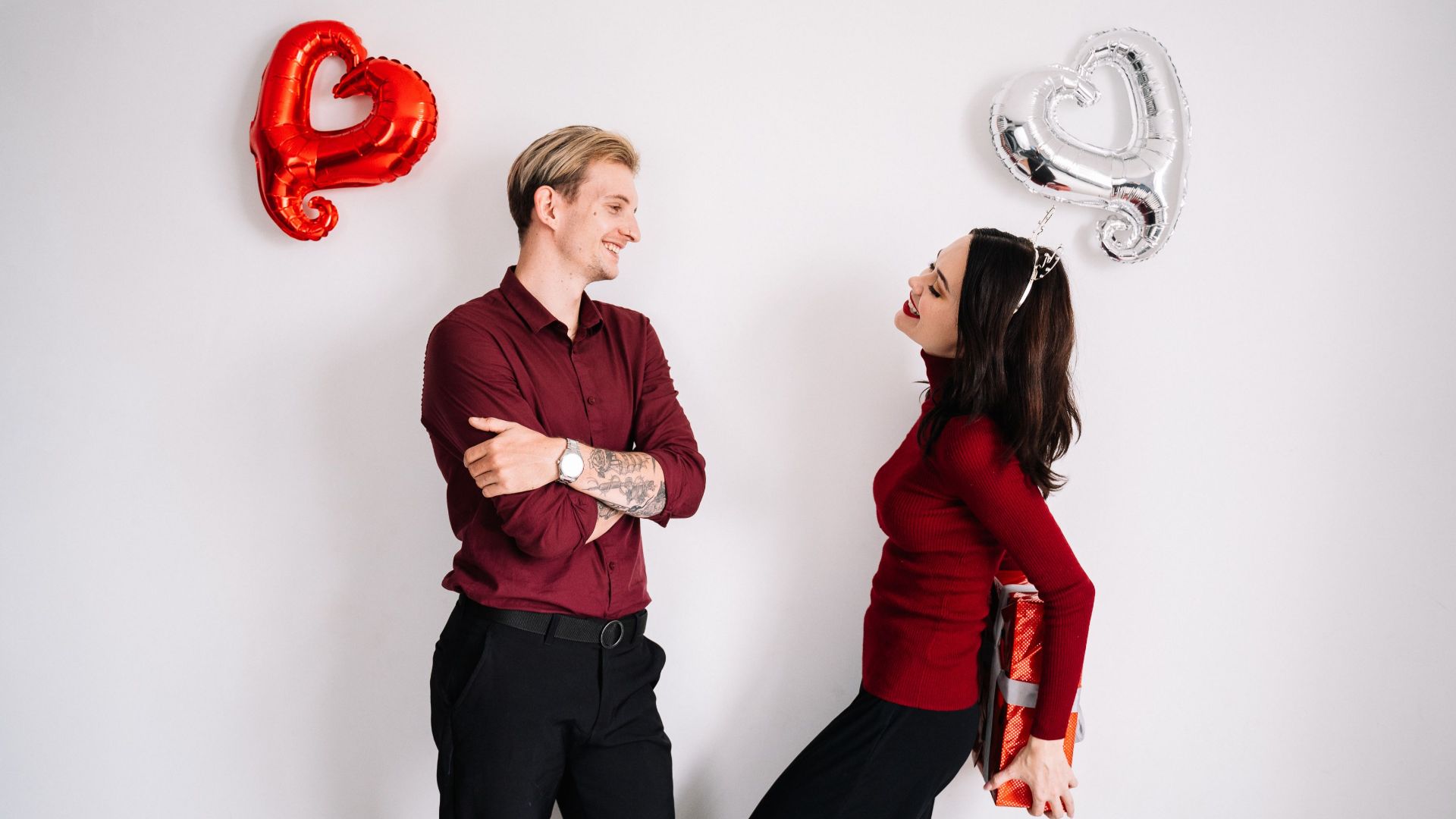 A happy couple celebrating love with heart-shaped balloons and a gift, indoors.