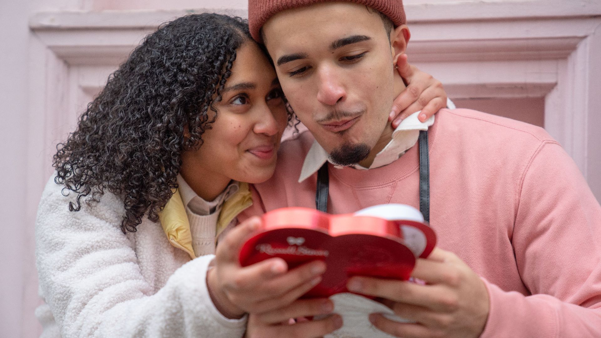 Joyful young ethnic couple embracing and eating yummy candies from heart shaped gift box during anniversary celebration