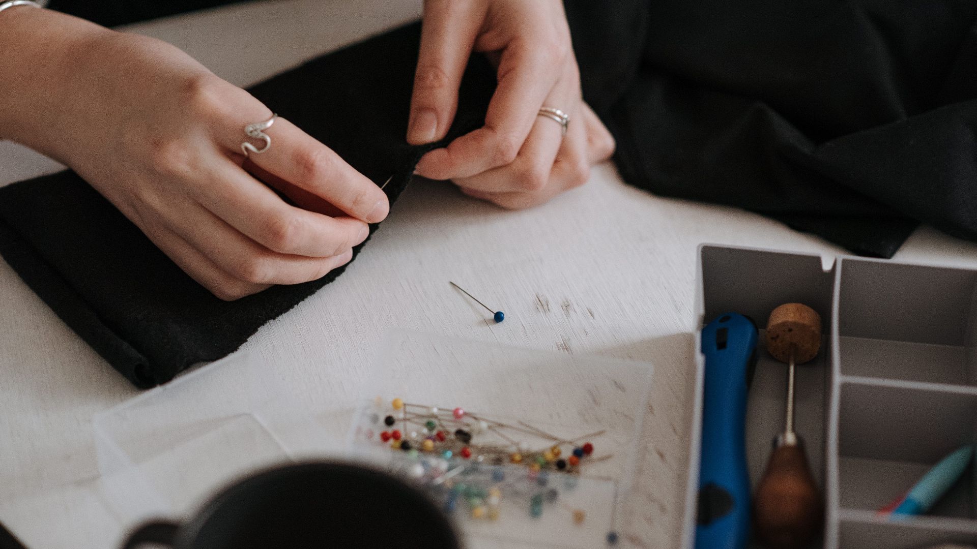 Unrecognizable female dressmaker using pins on black textile while sewing at table with container of professional instruments in modern studio