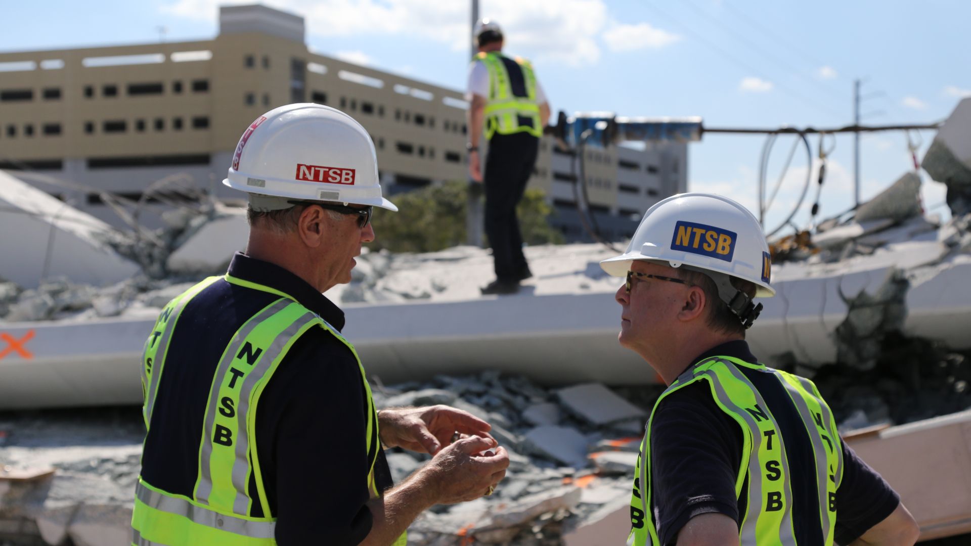 A member of the NTSB’s Go Team that is investigating Thursday’s collapse of a bridge on the FIU campus, examines debris while Investigator in Charge Robert Accetta briefs NTSB Chairman Robert Sumwalt on the status of the investigation. (NTSB Photo by Chri