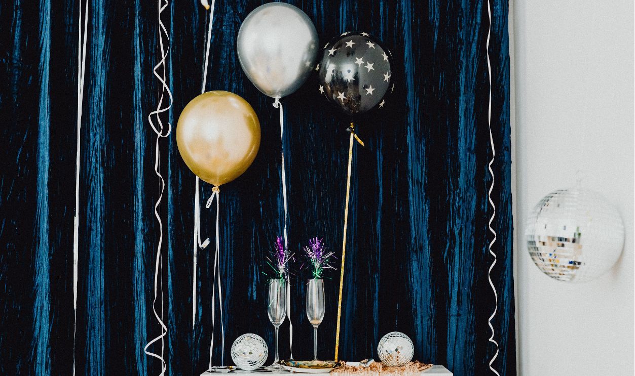 Stylish New Year Eve party decor featuring balloons, disco balls, and wineglasses against a blue curtain.