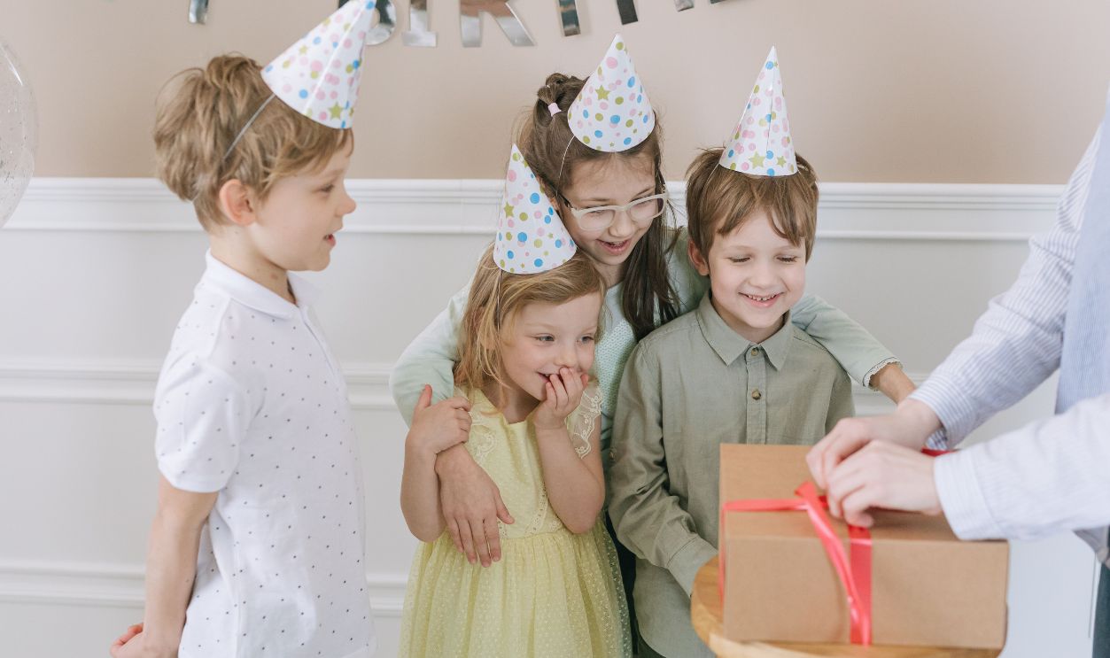 Happy children in party hats eagerly awaiting a gift at a birthday celebration indoors.