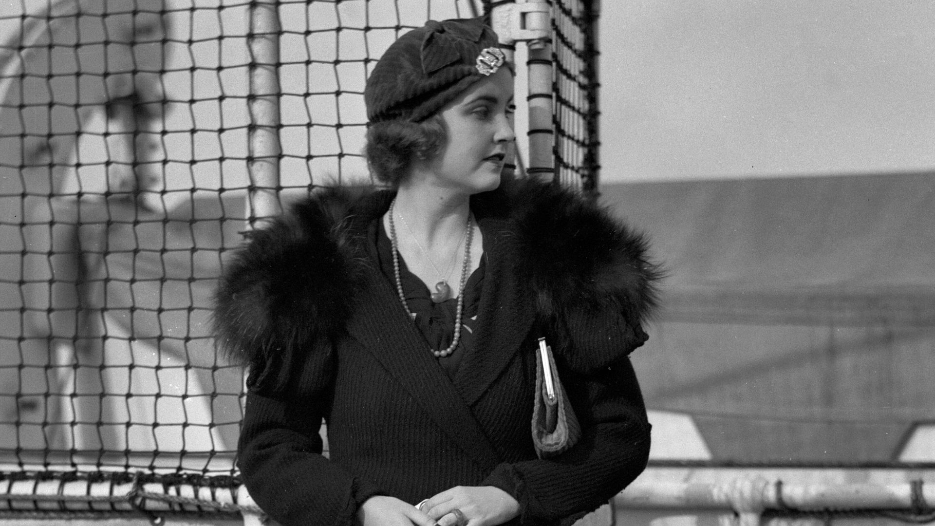 Barbara Hutton standing on a ship and looking to her left. She is holding a pair of white gloves in her hands.