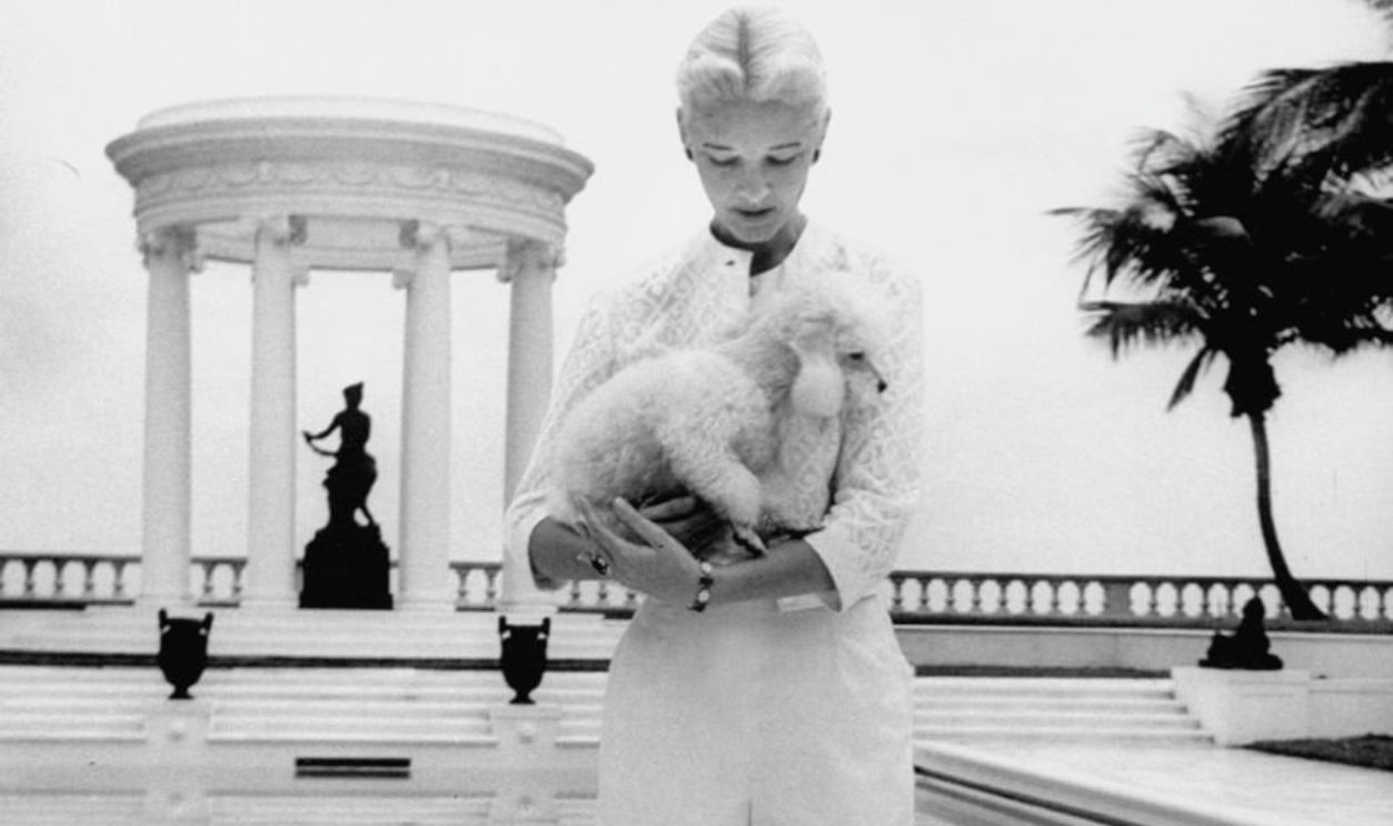 1956: CZ Guest, in white shorts, standing by her Palm Beach swimming pool, with white poodle, posing on black pool-side sphinx.