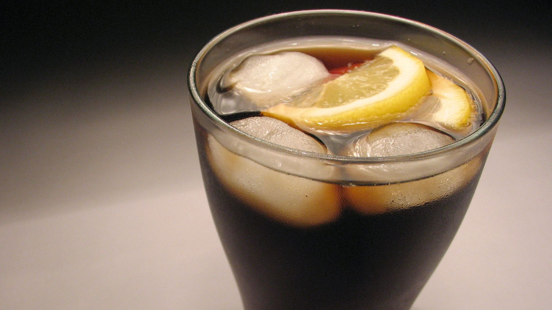 A glass of cold cola with three icecubes and two lemon pieces.