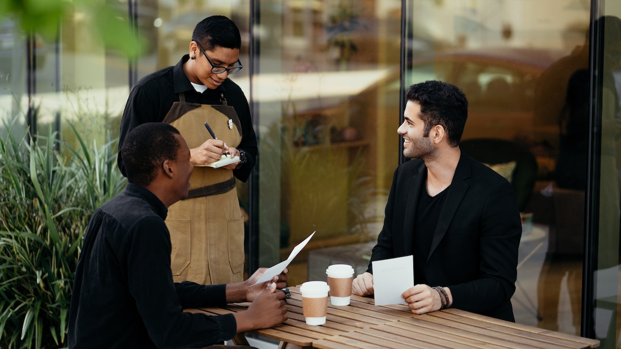 Waiter at restaurant is talking with two man seating at table.