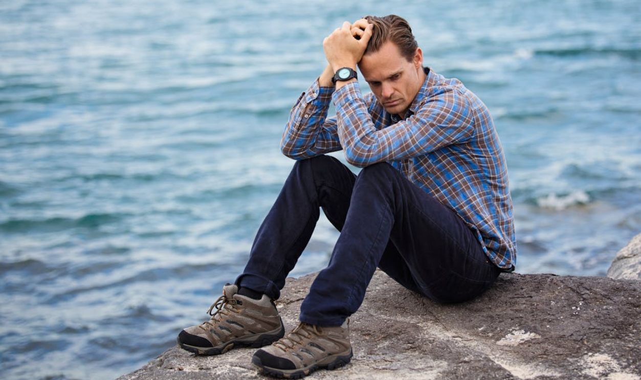 Man wearing blue and maroon plaid shirt sitting on a rock near a body of water