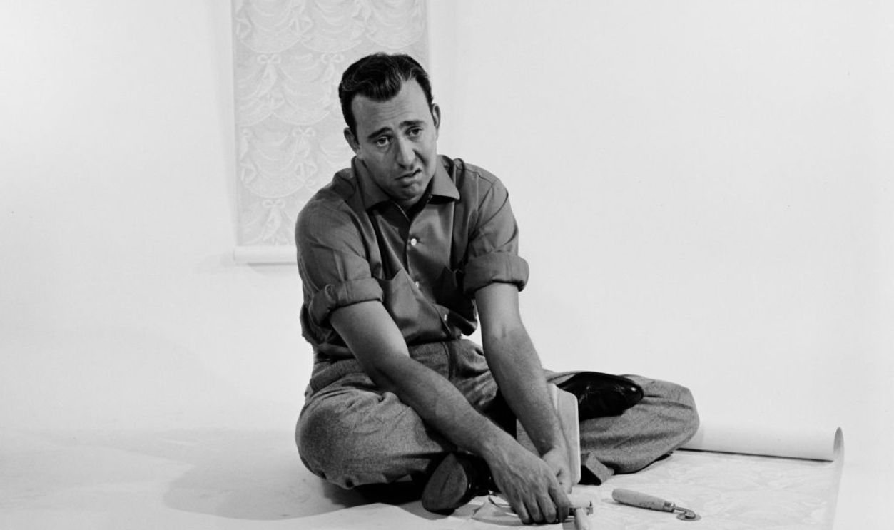 American comedian, actor and film director Carl Reiner, wearing a shirt with the sleeves rolled up, sits with a disgruntled facial expression, cross-legged before a length of wallpaper, against a white background during a studio portrait session, United States, 15th September 1955. 