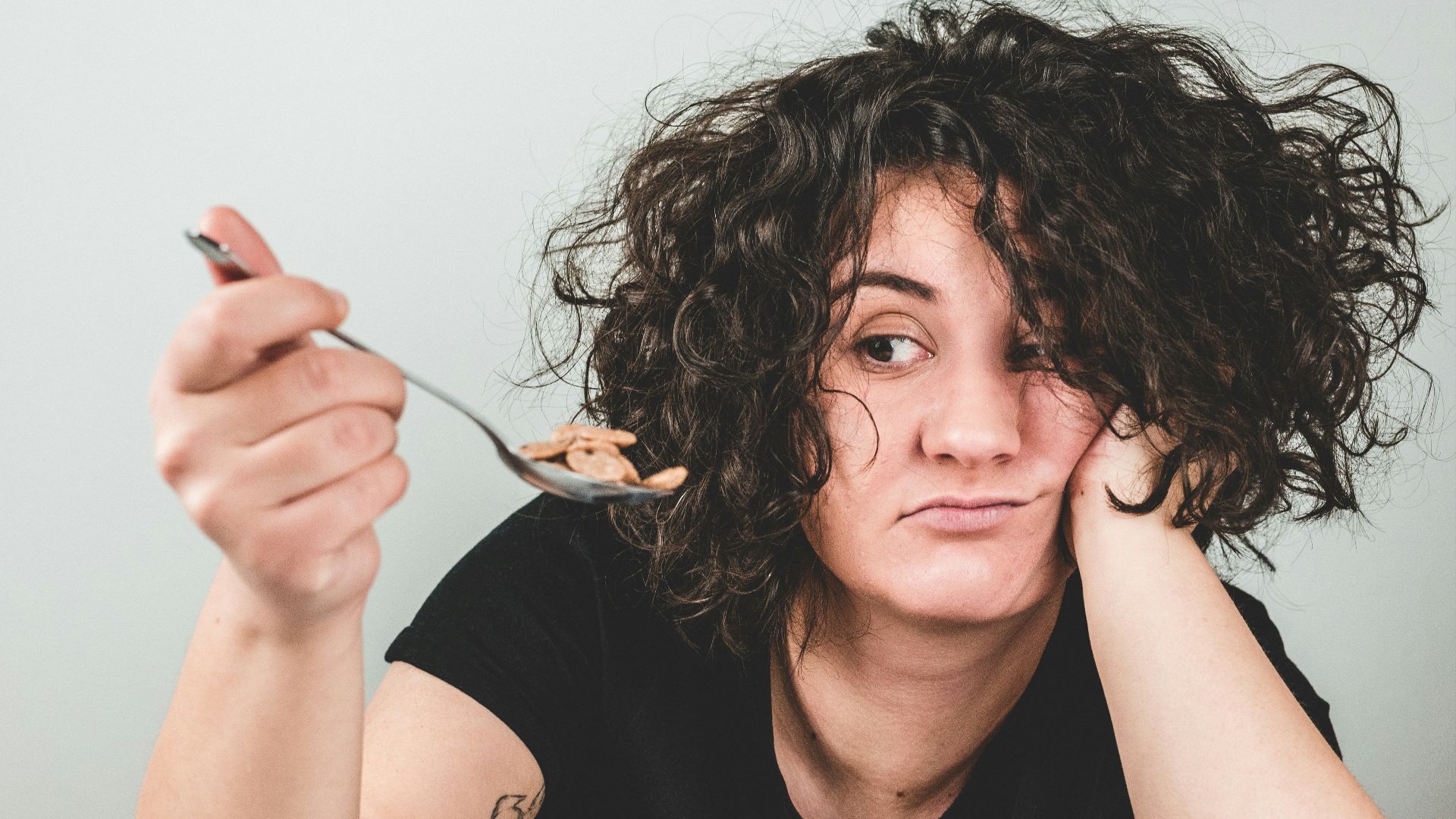woman with messy hair wearing black crew-neck t-shirt holding spoon with cereals on top