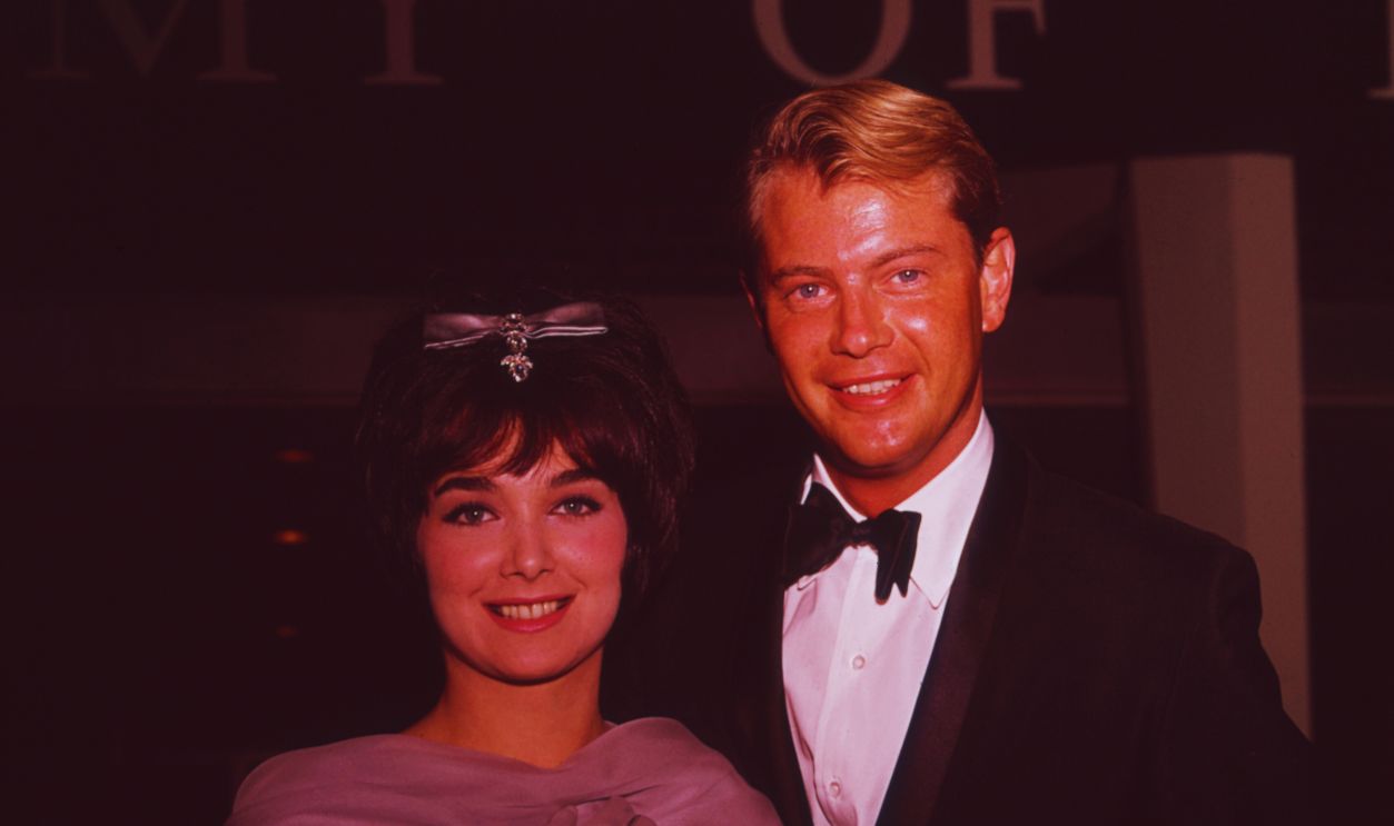Gettyimages - 3243609, Pleshette And Donahue 1964: American actor Suzanne Pleshette and her husband, actor Troy Donahue (1936 - 2001) smile as they arrive together at a formal event. She is holding her jewelled purse.