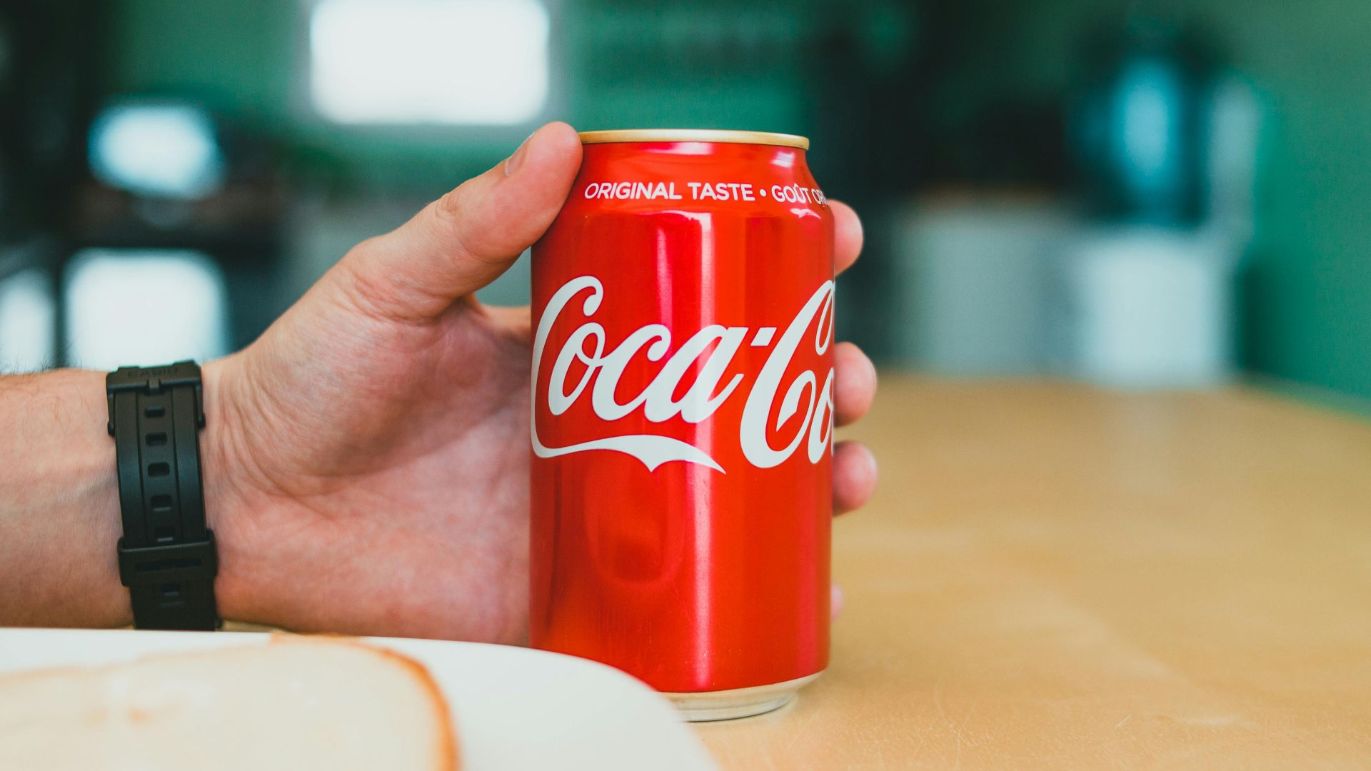 A person holding a red Coca-Cola can on a wooden table indoors.