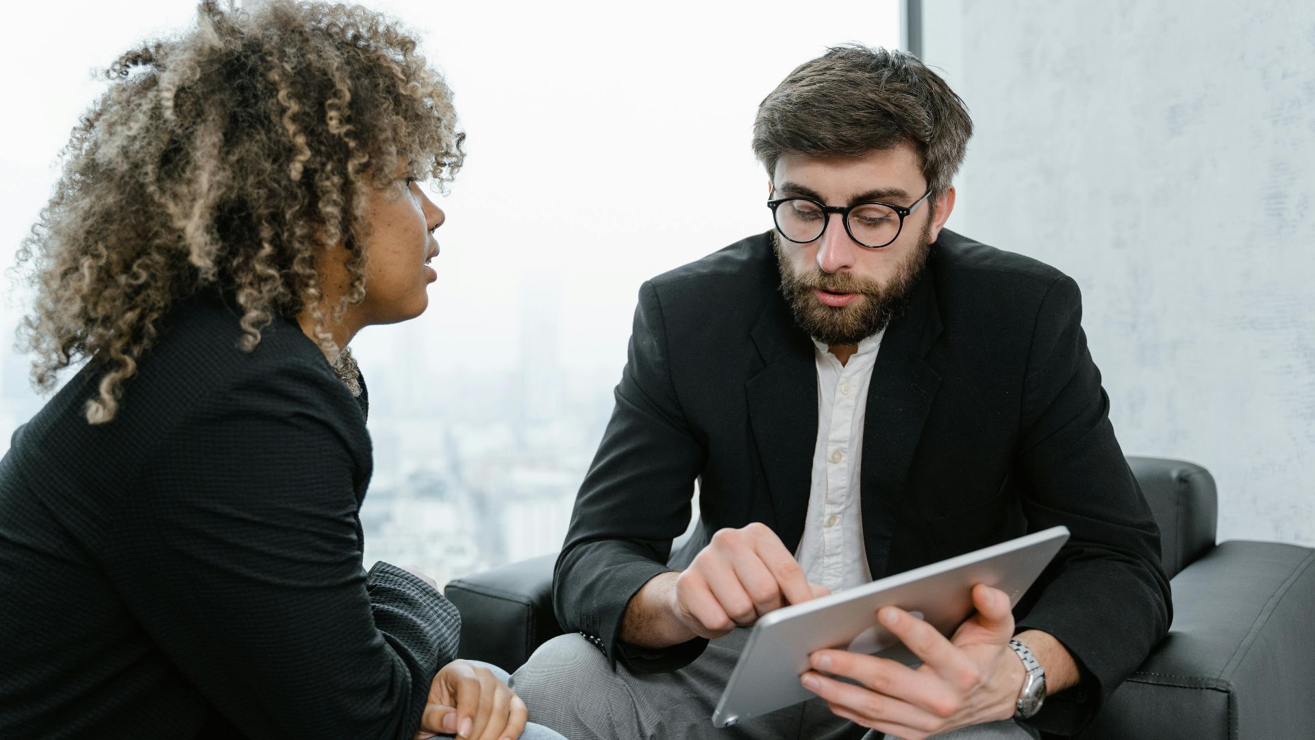 A focused business meeting between two professionals discussing data on a tablet indoors.