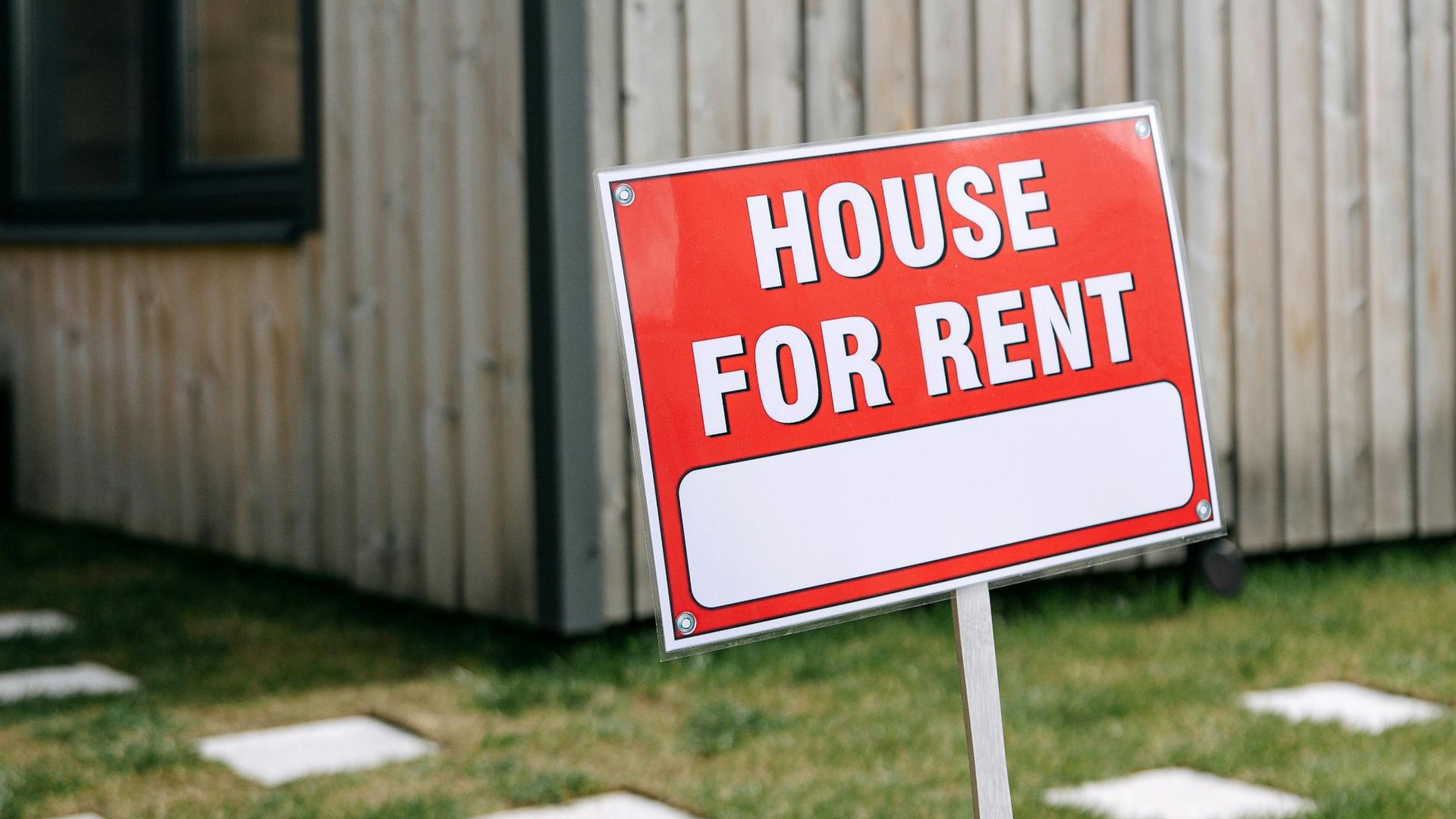 Red 'House for Rent' sign in front of a modern wooden house exterior.