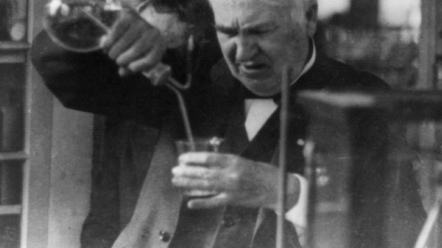 Thomas Edison, half-length portrait, facing left and looking down into glass, experimenting in his laboratory