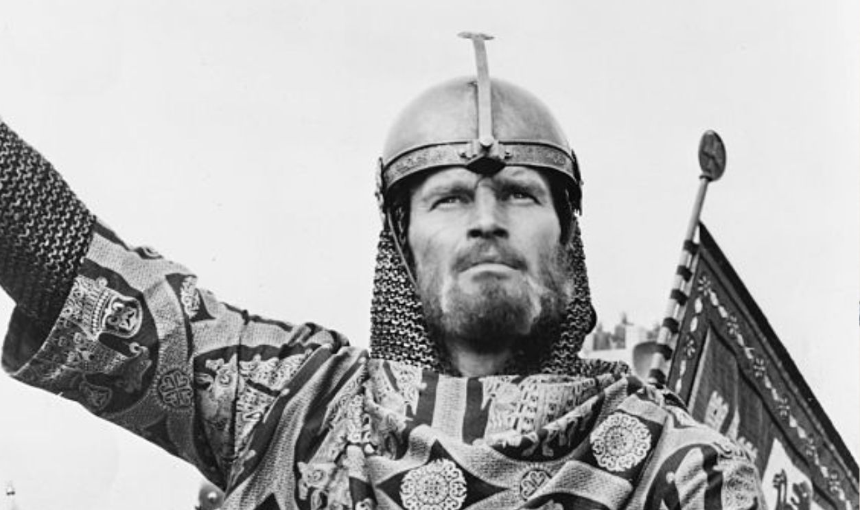 Charlton Heston in a still from the film El Cid. He is riding a horse, at the head of an army, holding a wooden lance.