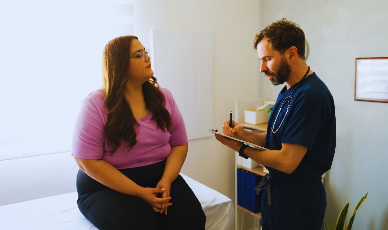 Plus size woman sitting on examination table while talking to doctor taking notes on clipboard during medical consultation in doctor's office