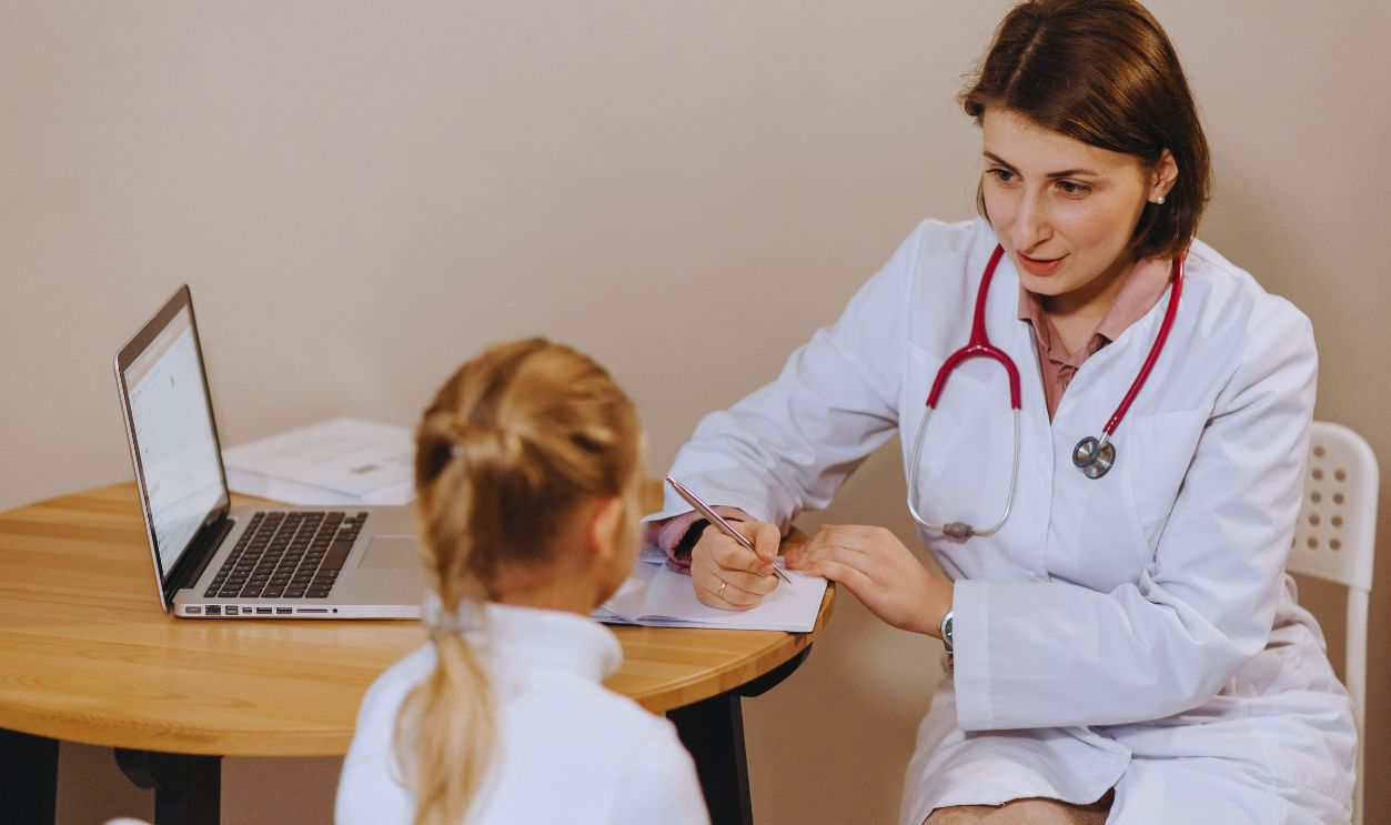 Woman in White Lab Coat Listening to a Girl and Writing Down Notes