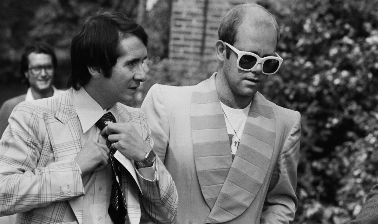 Getty Images - 1168937752 - English singer, songwriter, pianist, and composer Elton John and his manager John Reid on their way to a luncheon in honour of Buddy Holly at the Orangery in Holland Park, London.