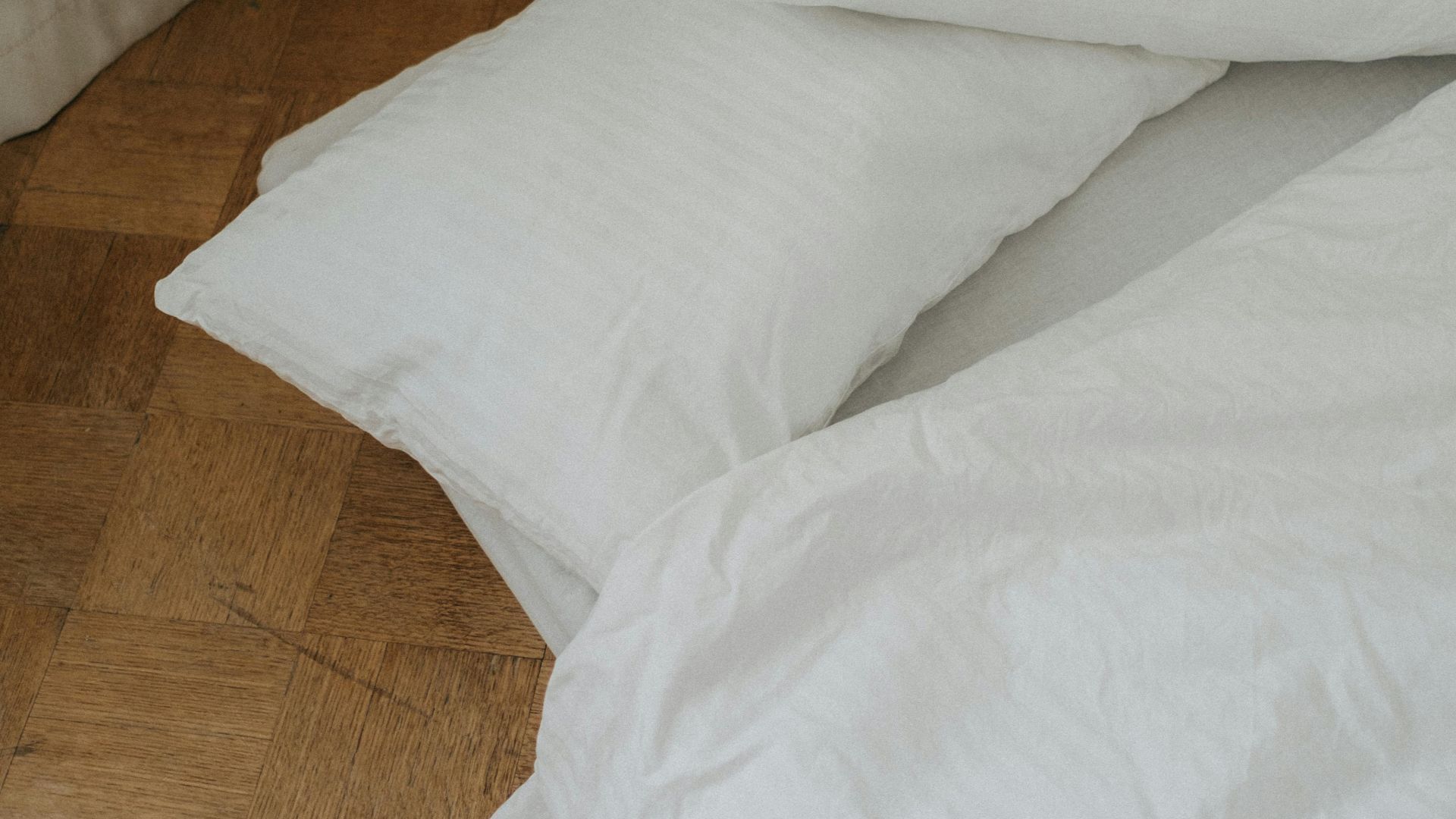 A simple minimalist setup with white bedding and pillows on a wooden floor, emphasizing simplicity and calm.