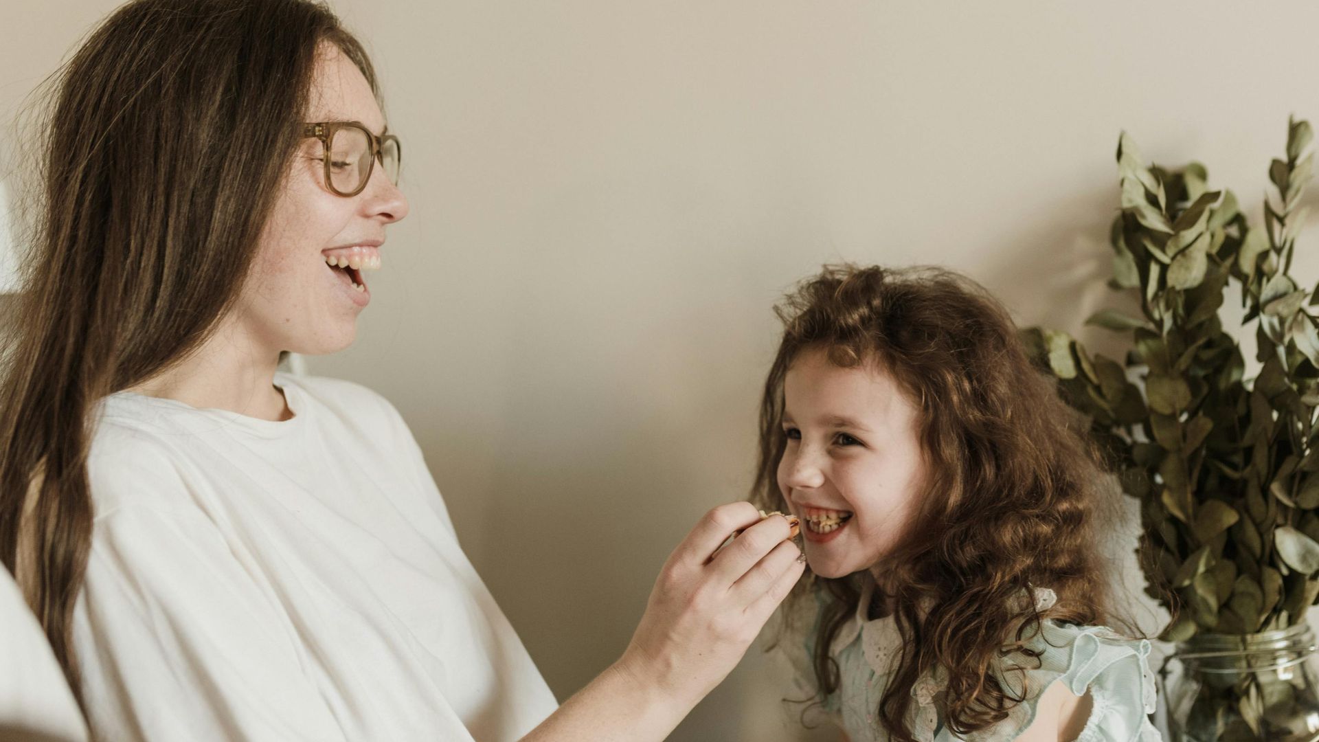 A mother joyfully feeds her smiling daughter a cookie inside their cozy home.