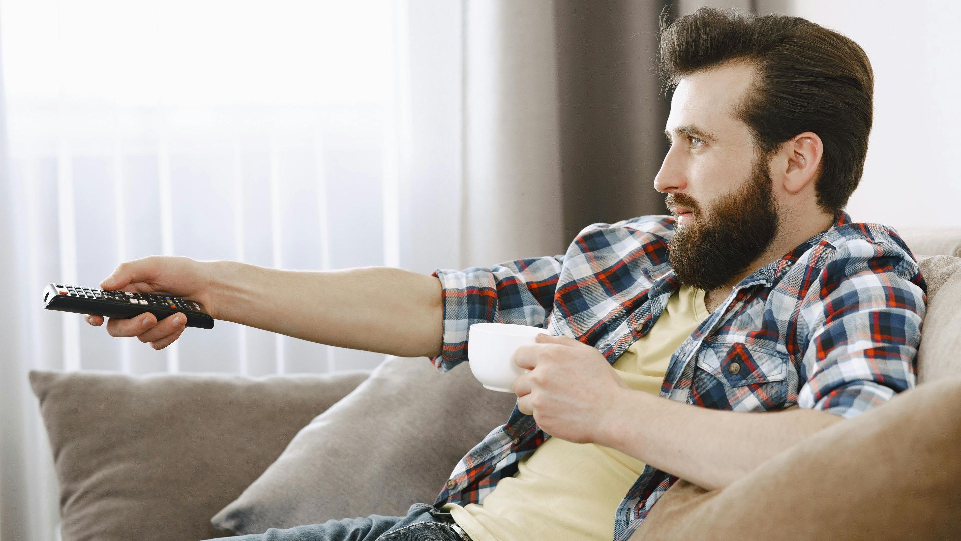 Bearded man in casual wear relaxing on a sofa, enjoying coffee while watching TV indoors.