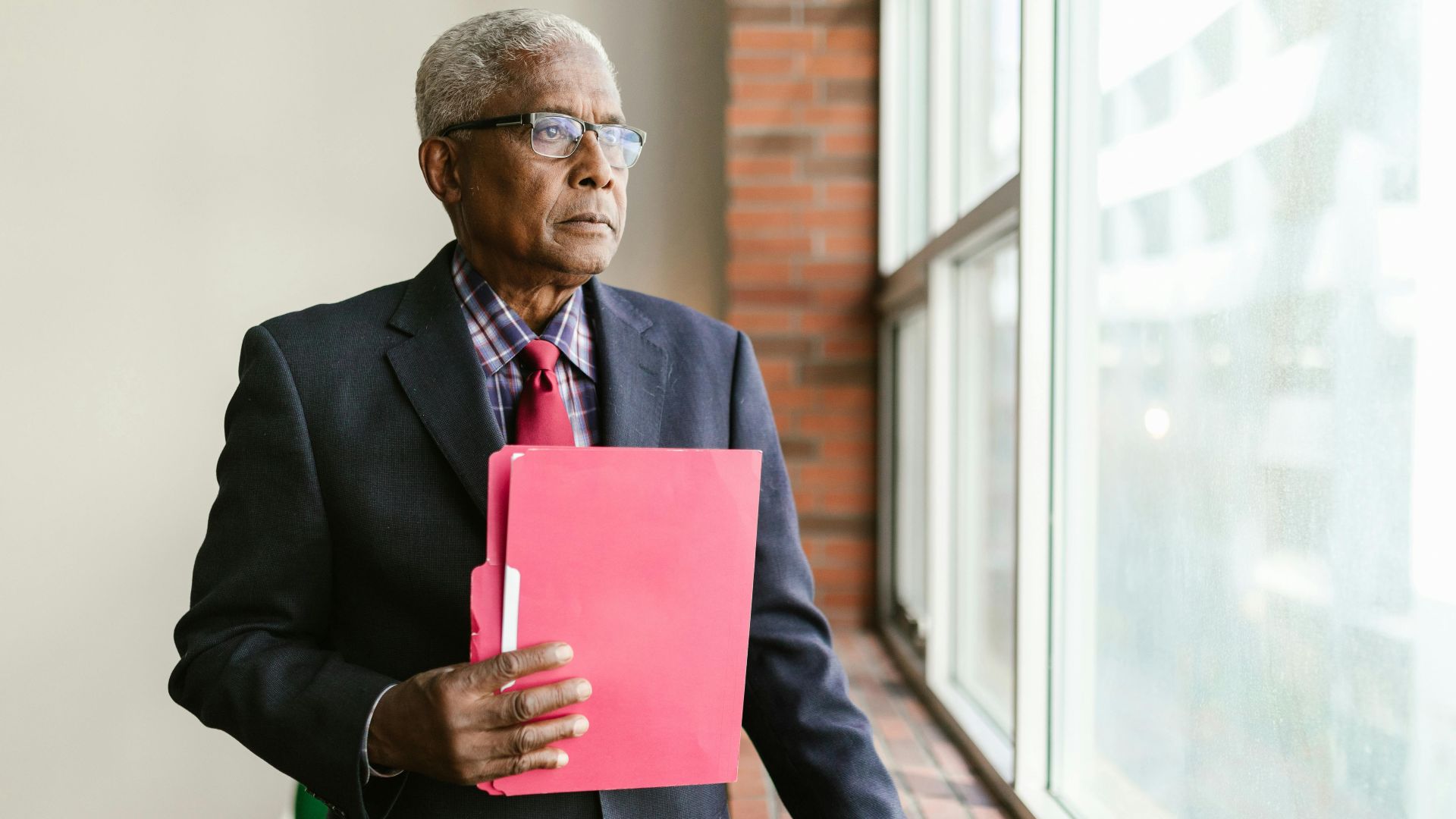 Mature man in suit and glasses holding a folder, gazing out the window.