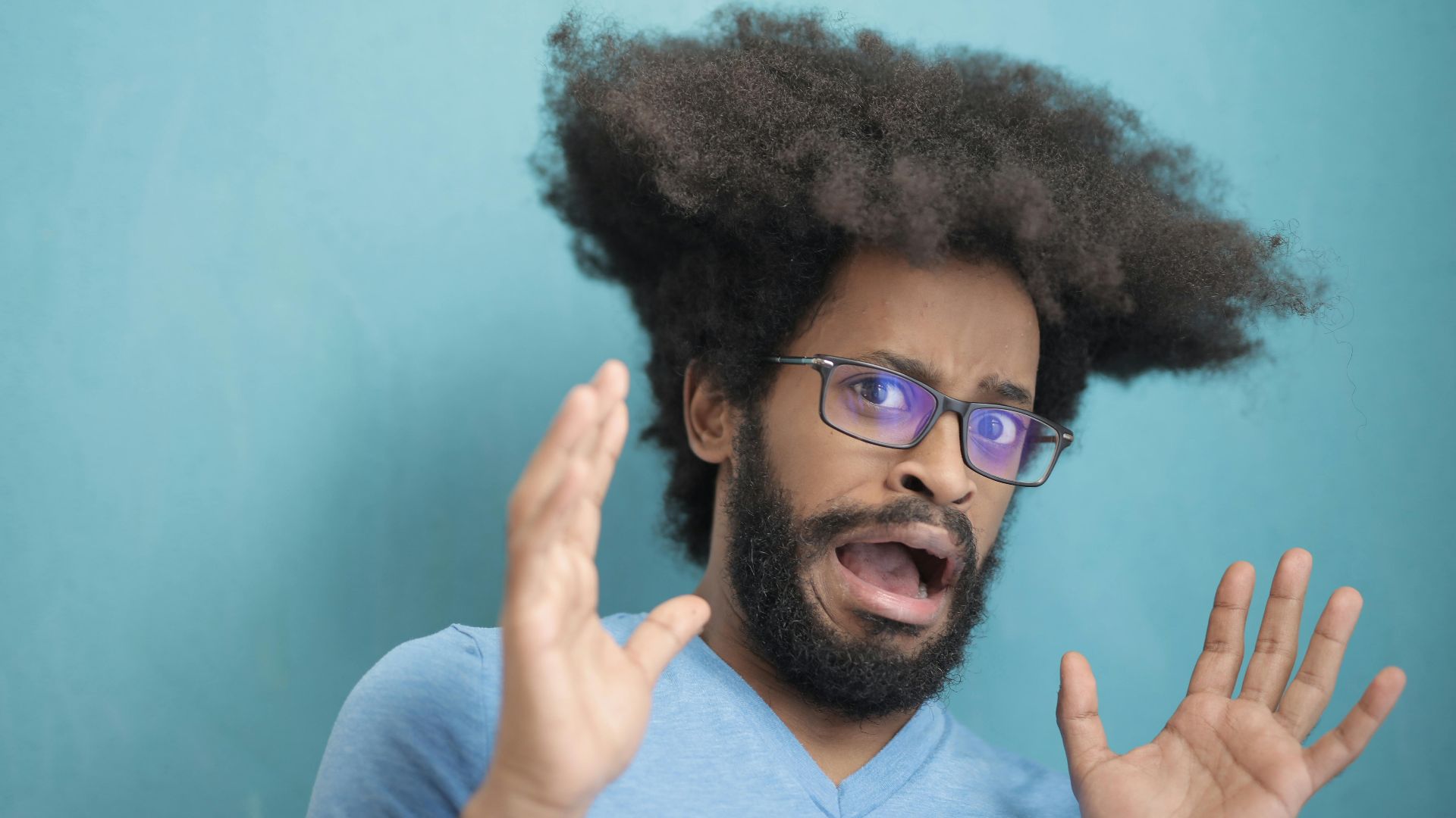 Portrait of a surprised man with afro hair and glasses, expressing shock with a blue background.