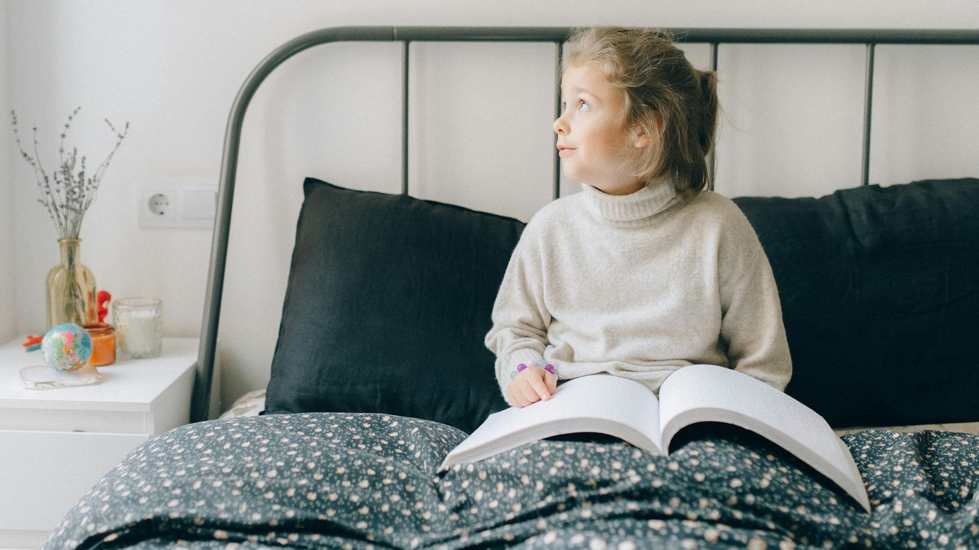 A young girl reading a book on a bed in a cozy bedroom setting, looking thoughtful.