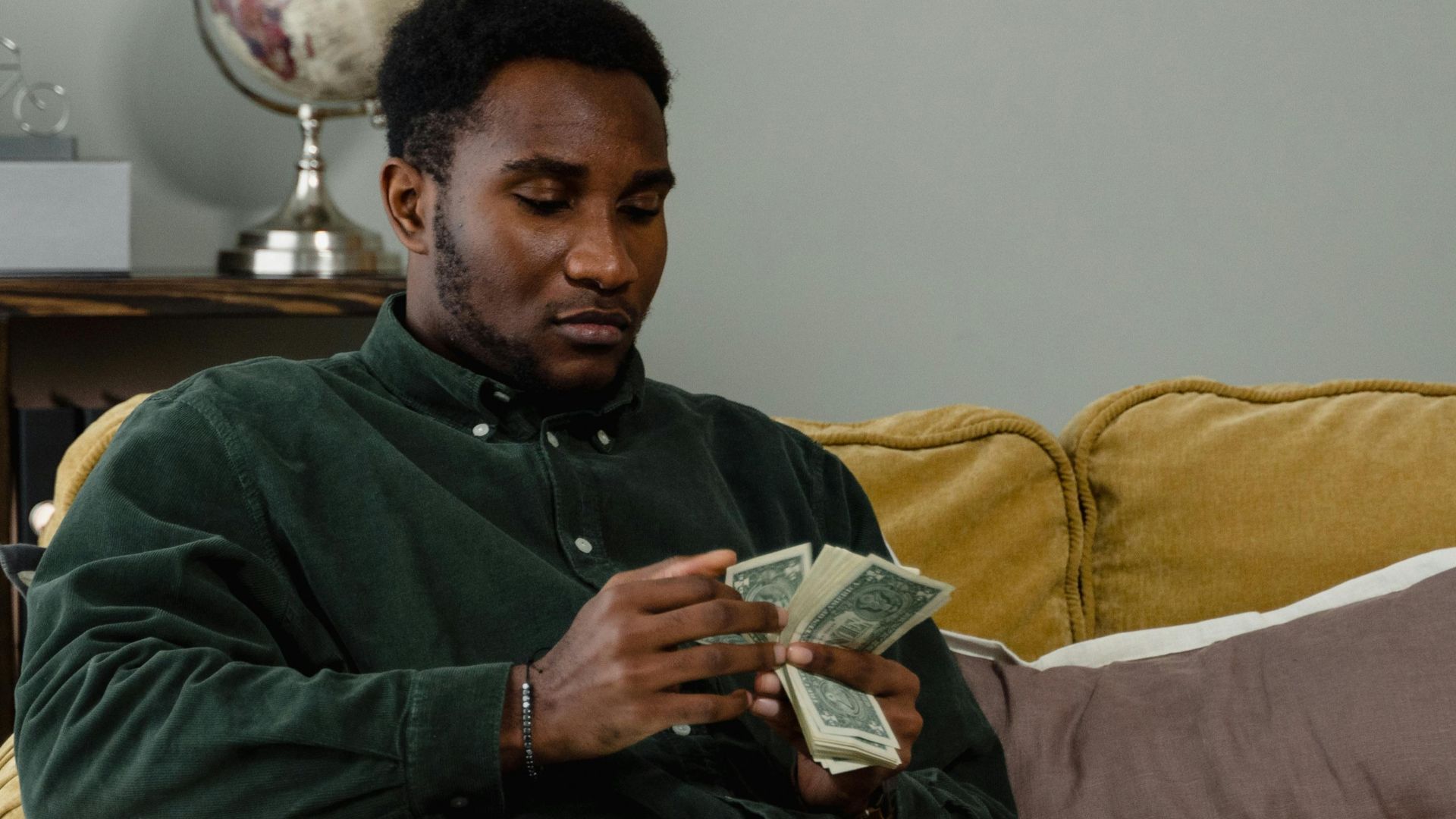 A man sitting on a couch counting cash notes in a cozy indoor setting with a globe in the background.