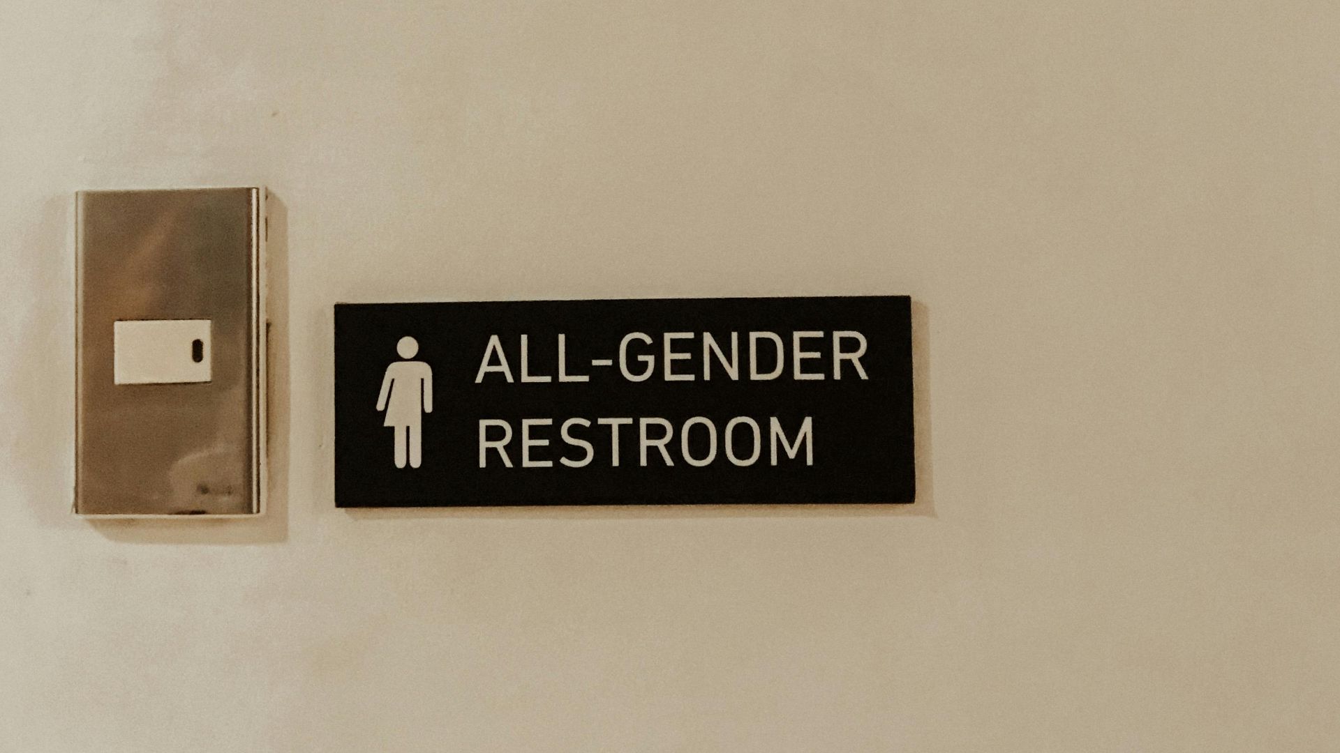 A minimalist sign for an all-gender restroom beside a light switch on a plain wall.